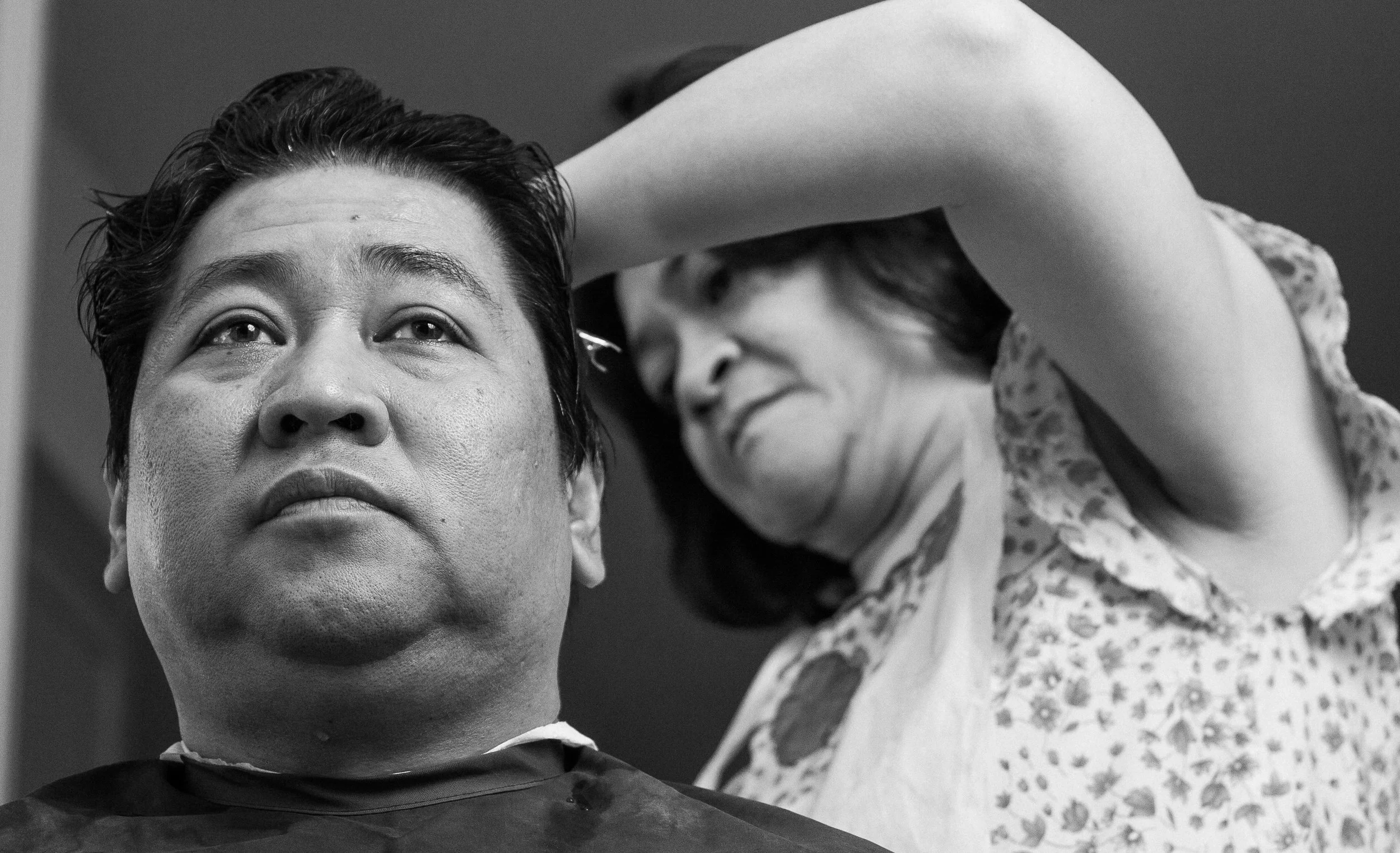 A woman with short hair is in a barbershop chair, having her hair cut or styled by a hairdresser with short hair, wearing a patterned shirt.