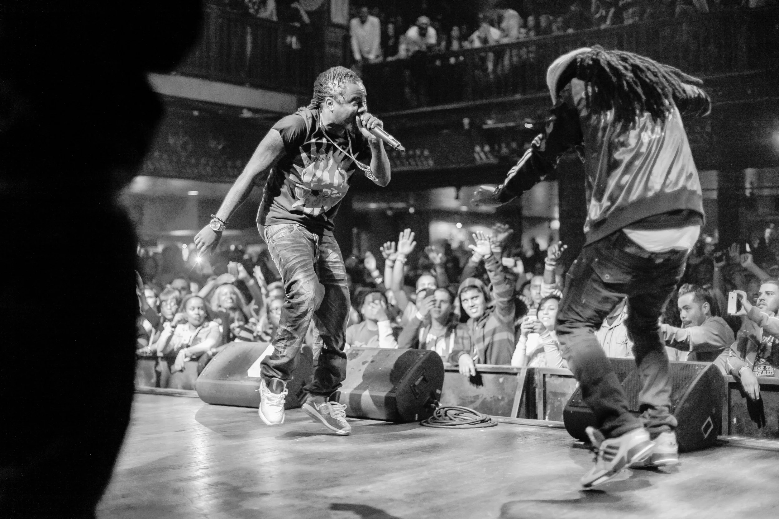 Black and white photo of a live concert. The rapper, Wale, holding a microphone, is performing on stage. Audience members are visible in the foreground, reaching towards the stage, with some taking photos. 