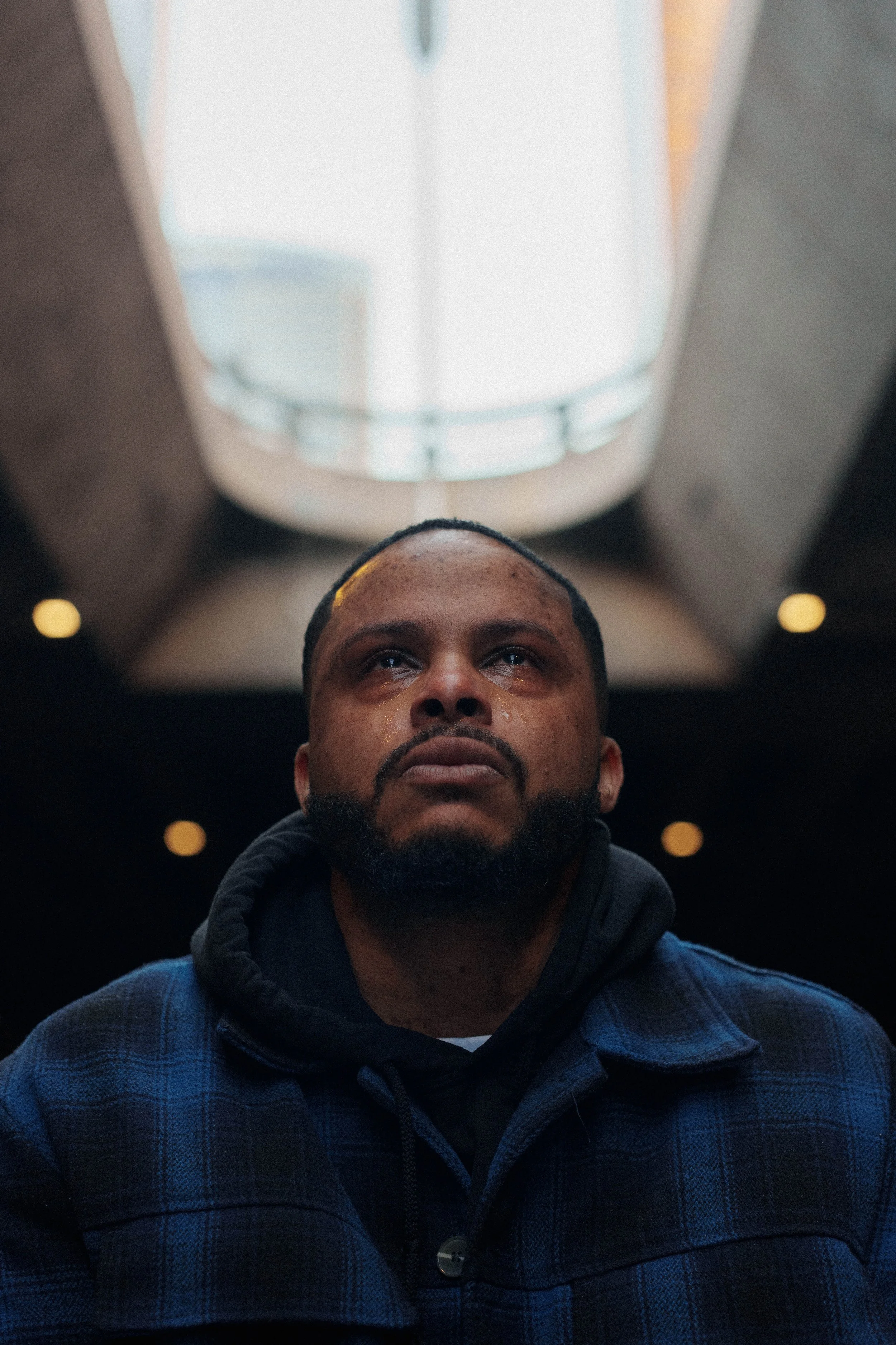 A man with a serious expression, looking upward, in an indoor setting with a ceiling light above his head, wearing a dark hoodie and a blue checkered jacket.