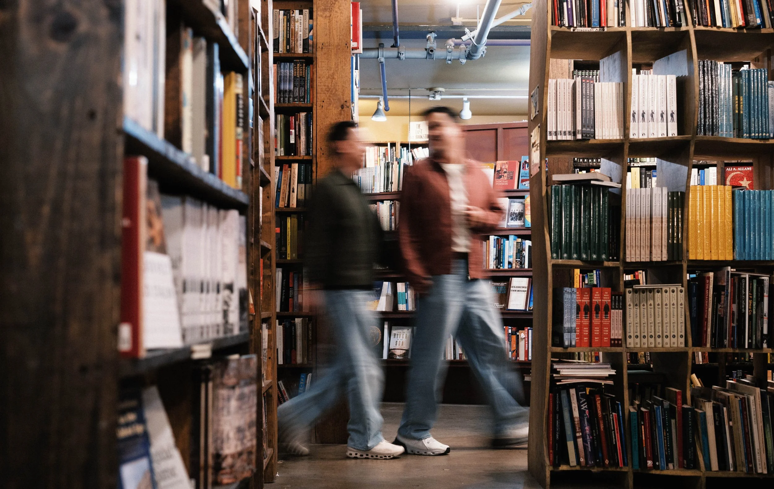 Two people walking past bookshelves in a bookstore or library, with both of their faces blurred, wearing casual clothing and sneakers.