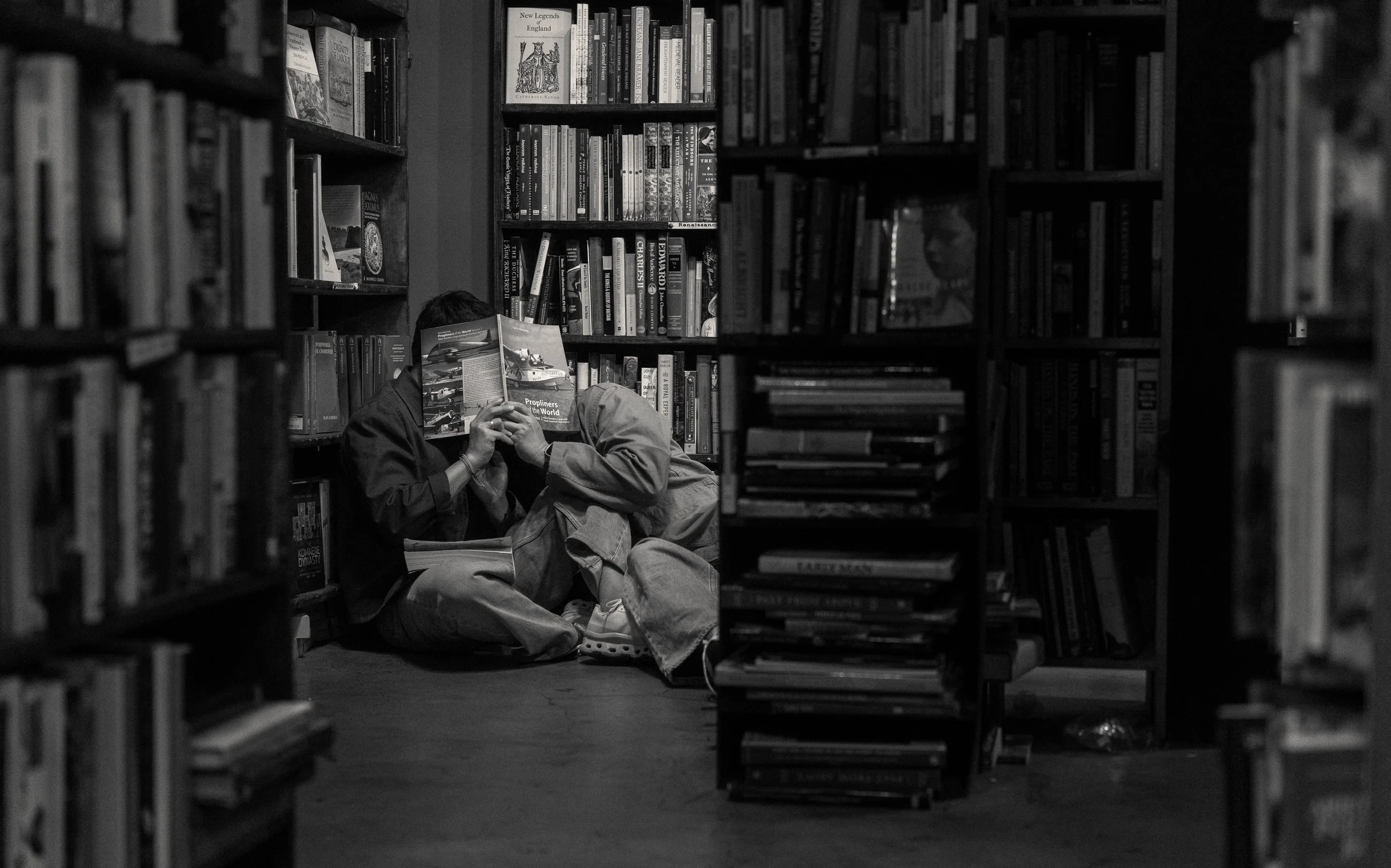 Two people sitting on the floor of a library aisle, reading a book together.