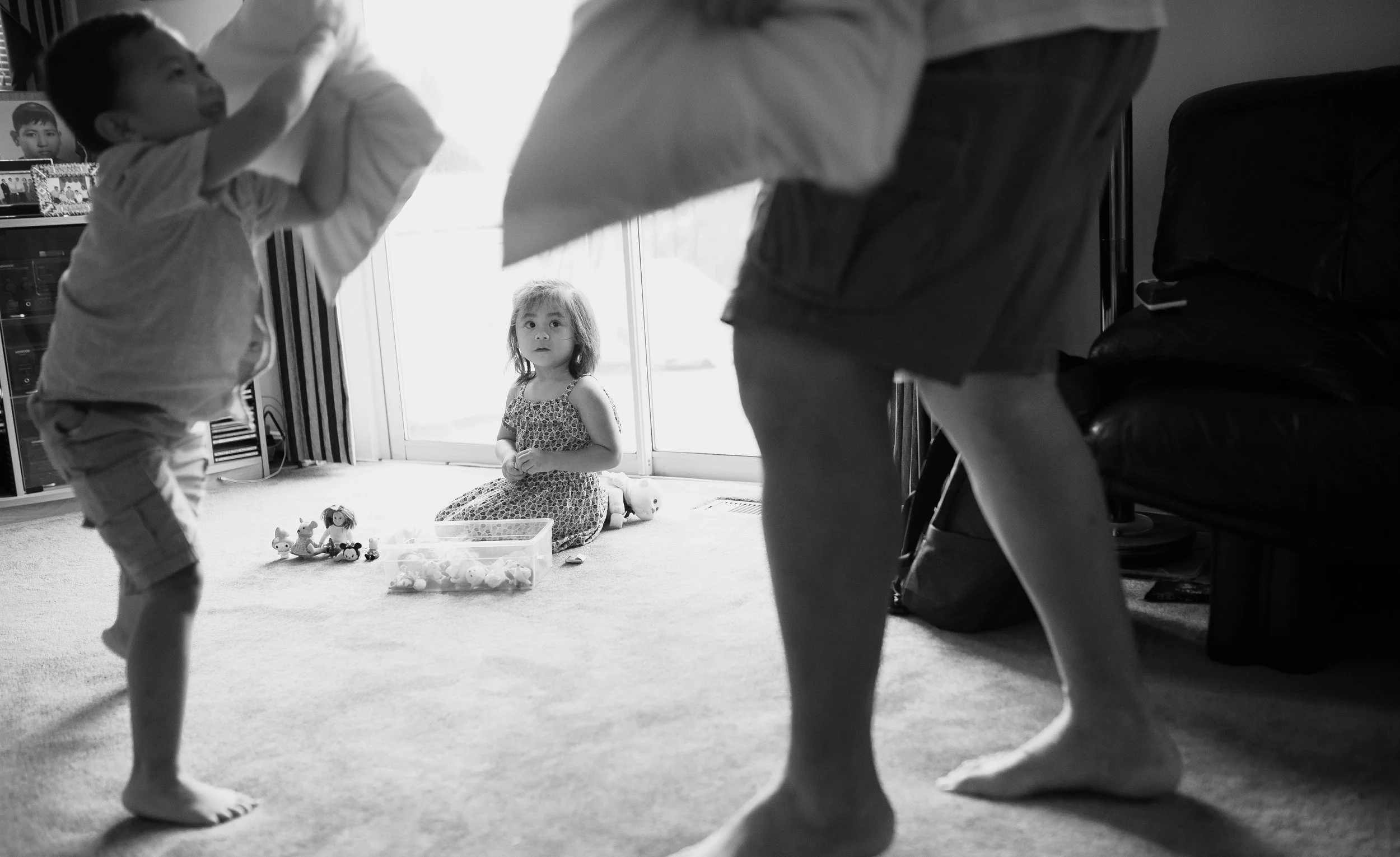 A young girl kneeling on the carpet in a room, surrounded by toys, looking at two children who are playing in the foreground. The scene is lit by natural light coming through a glass door behind her.