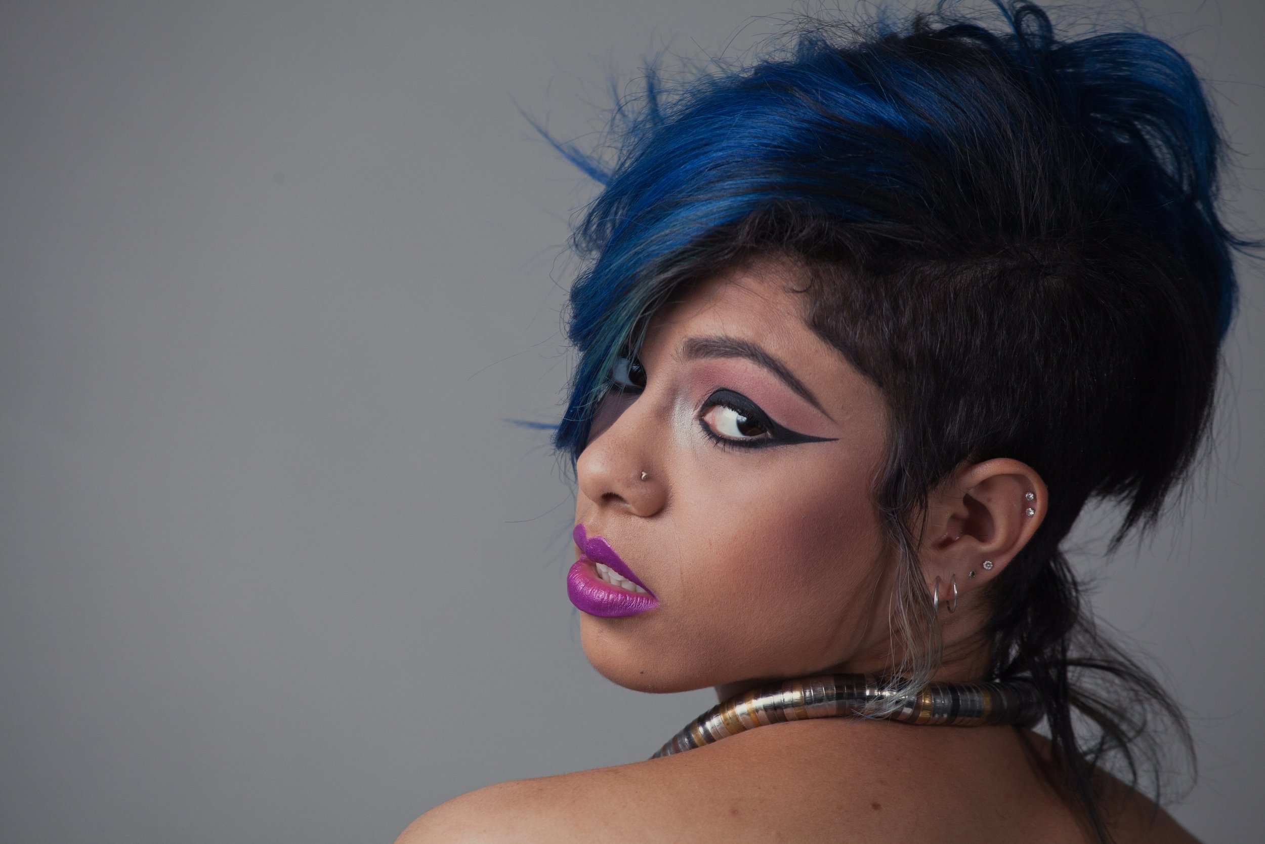 Close-up of a woman with short black hair with blue highlights, bold makeup including pink lipstick and winged eyeliner, wearing earrings and a metallic necklace, and looking over her shoulder against a plain background.