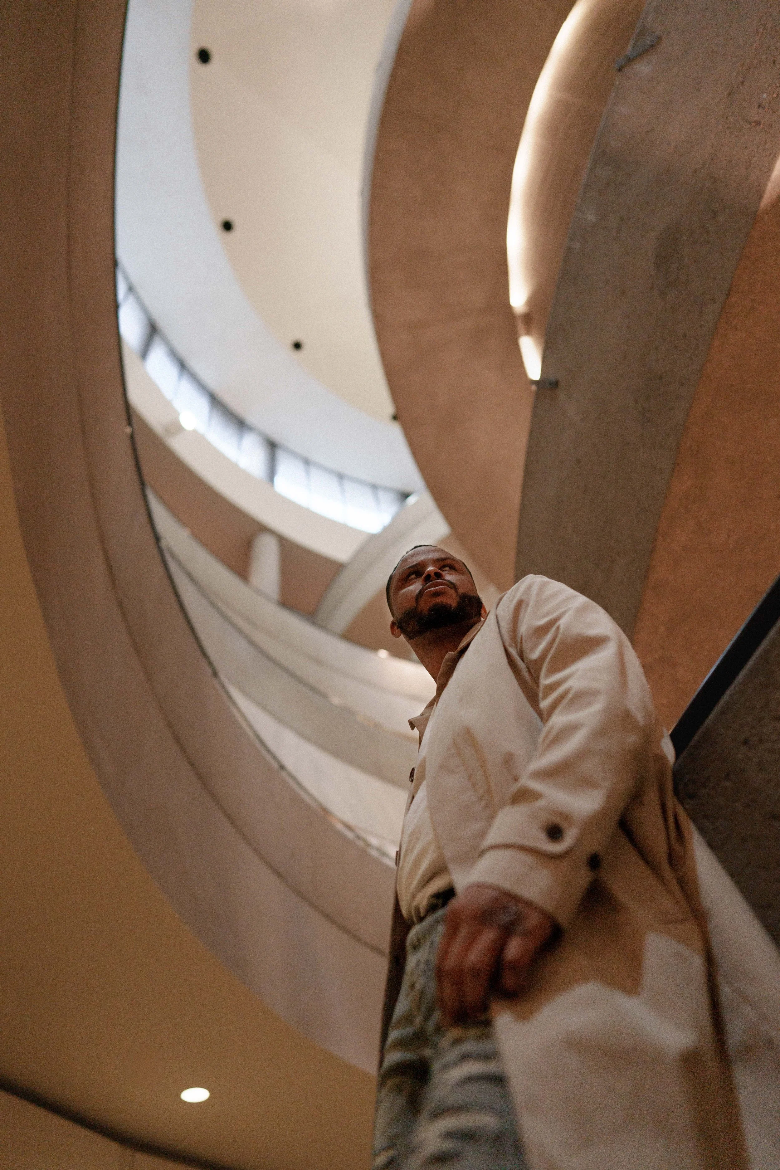 A man standing on an escalator in a modern indoor space, possibly a shopping mall or office building, with a curved ceiling and windows letting in natural light.