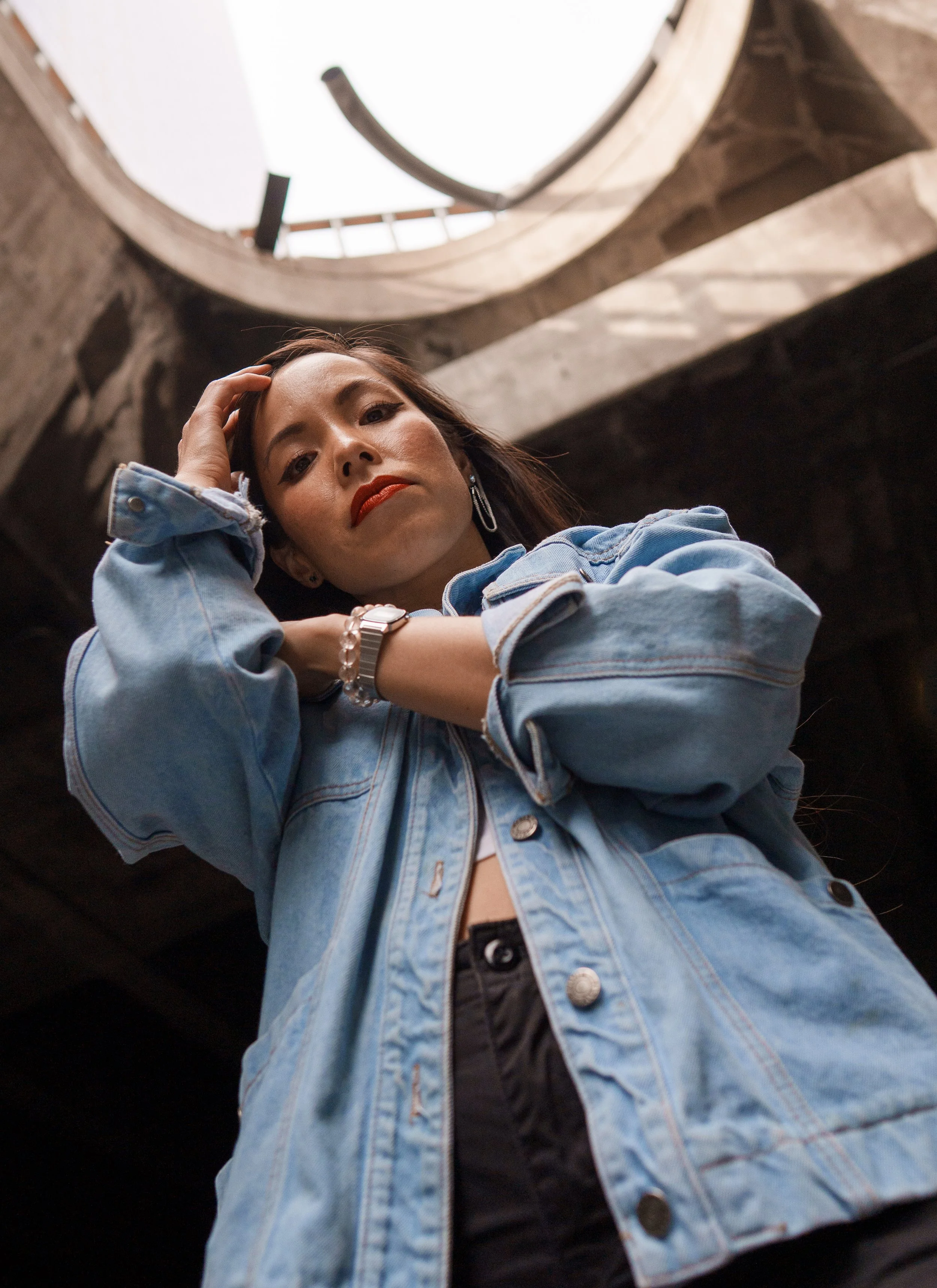 A woman with red lipstick and earrings looking down, wearing a denim jacket and black top, posing in front of a circular opening or window in a dimly lit indoor space.