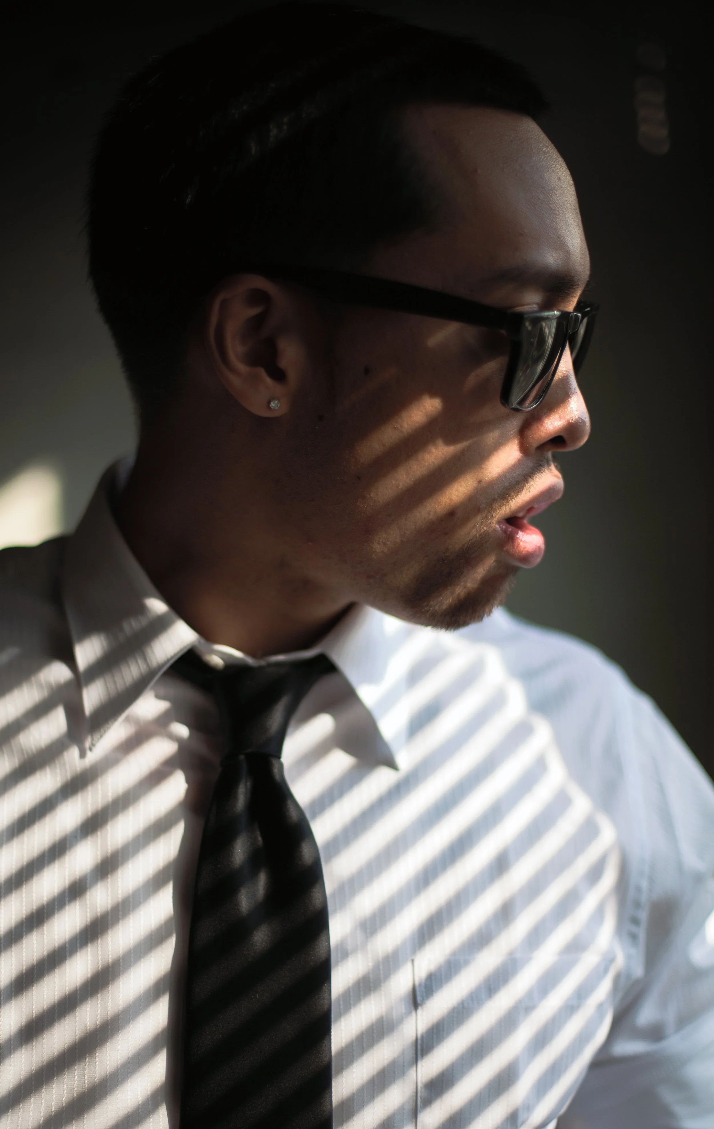 Profile of a young man wearing glasses, white dress shirt, black tie, and earrings, with light casting horizontal shadows across his face and clothing.