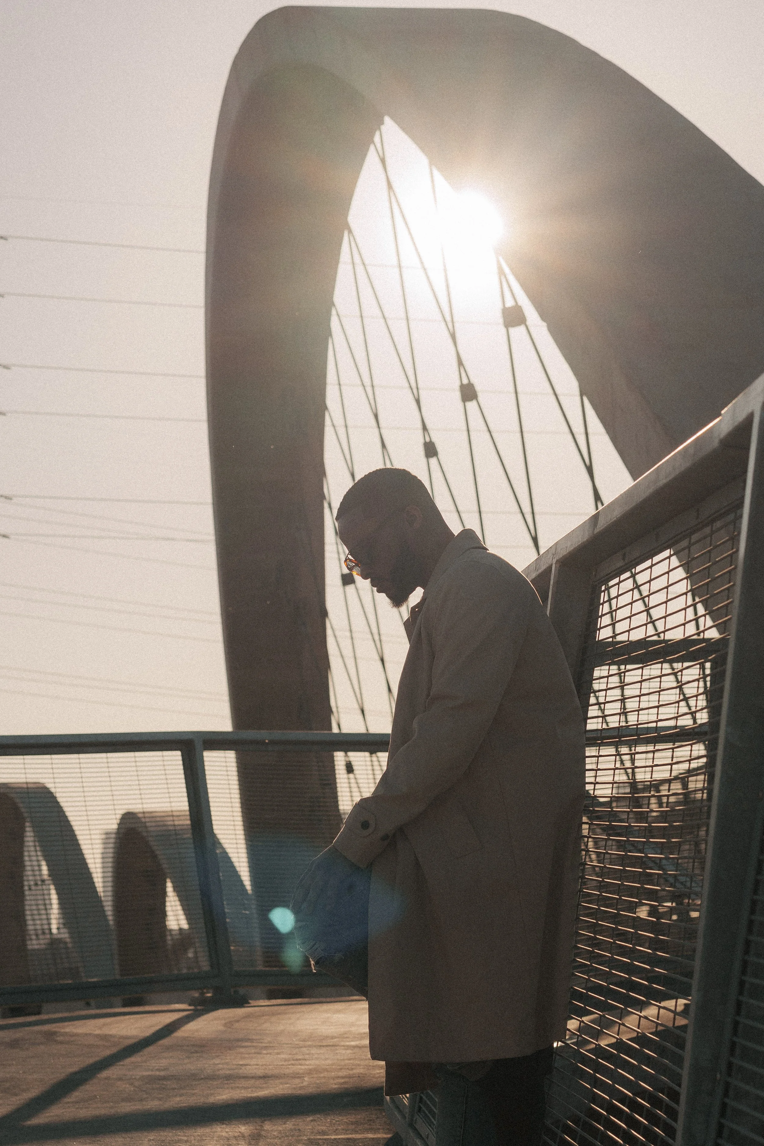A person wearing glasses and a beige trench coat stands on a rooftop or balcony, looking down, with the sun shining behind a modern architectural structure with curved and linear elements, possibly a bridge or a building. The person is in profile, wi