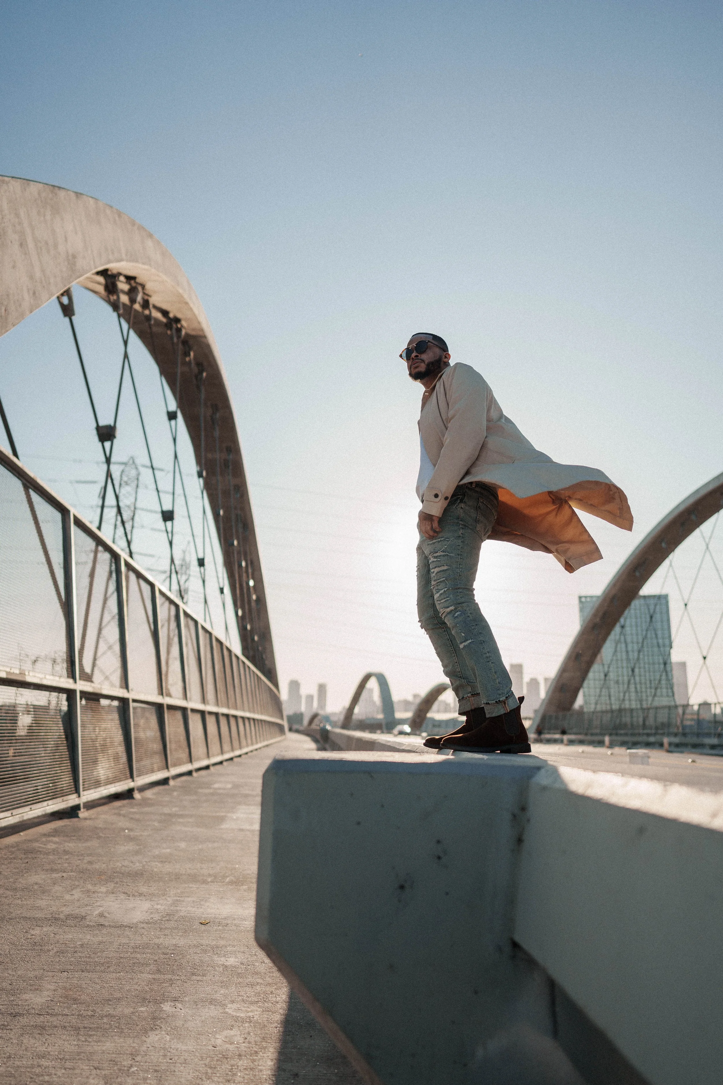 A man wearing sunglasses, a beige trench coat, and ripped jeans skateboarding on a city bridge during sunset with a modern city skyline in the background.