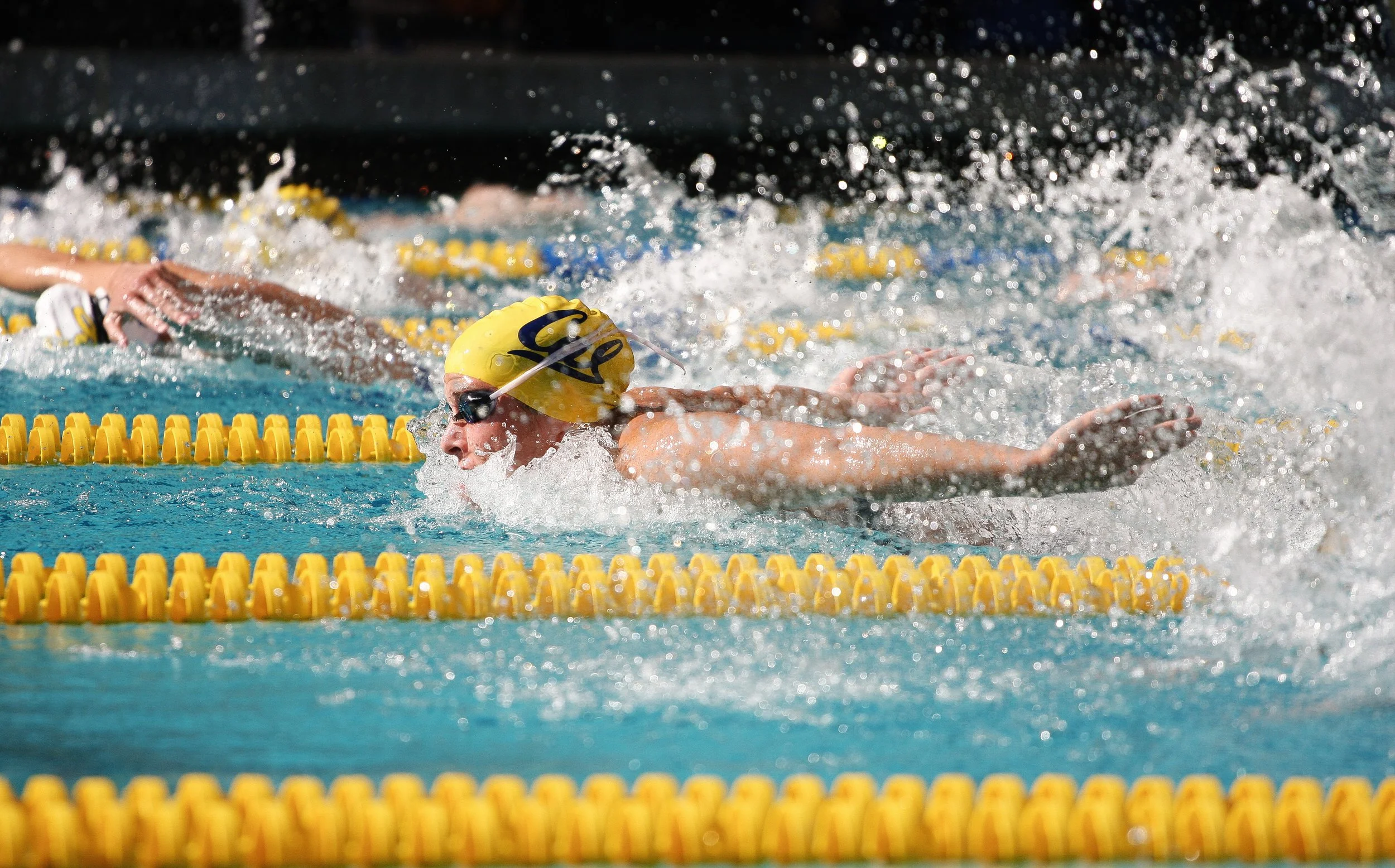 A swimmer in a yellow cap is swimming freestyle in a pool, with water splashing around and lanes marked by yellow lane dividers.