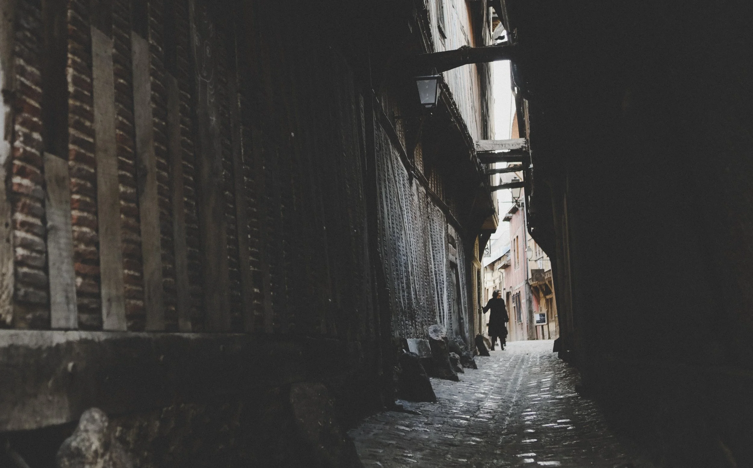 A narrow street viewed from inside a dark alley, revealing older buildings on either side. Two people are walking in the distance with umbrellas, suggesting rainy weather.