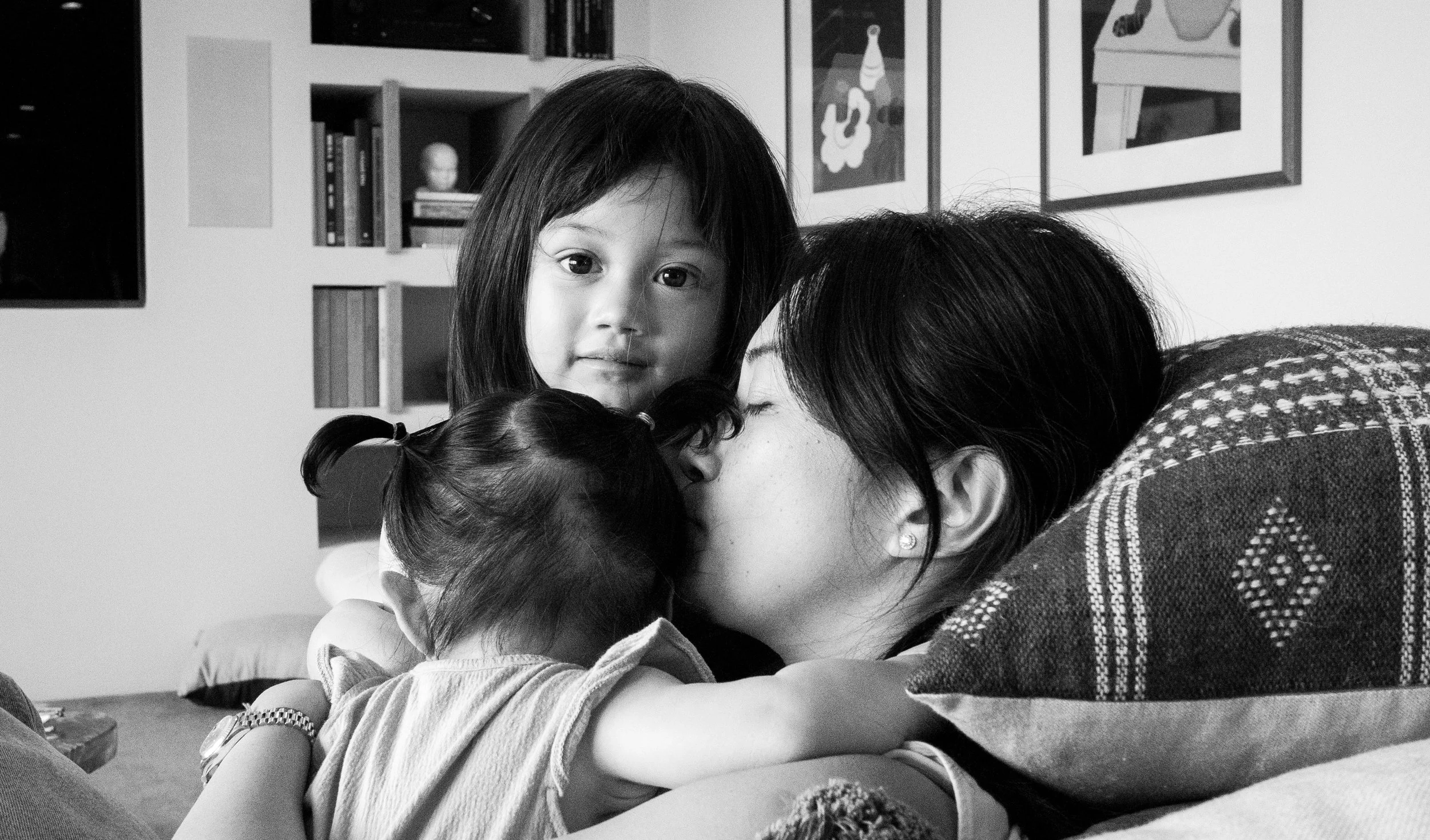 A mother is hugging and kissing her two children, one with pigtails and another with long hair looking into the camera, in a cozy room with art and books on the wall.