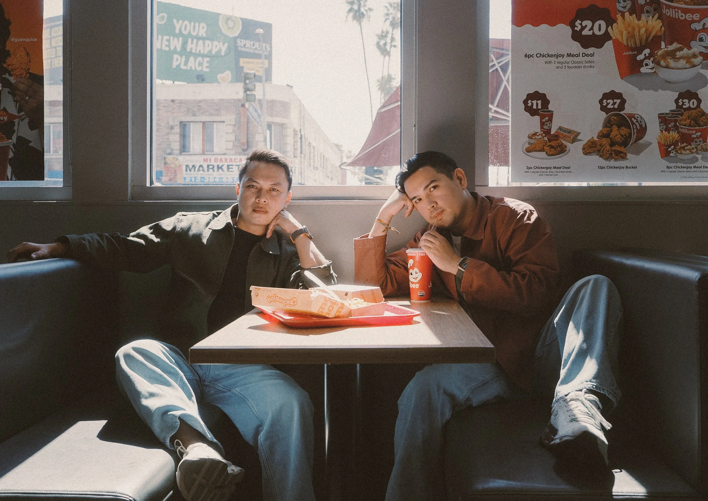 Two young men are sitting at a table in a fast food restaurant, eating and posing for a photo. They are sitting next to a large window with signs and buildings visible outside.