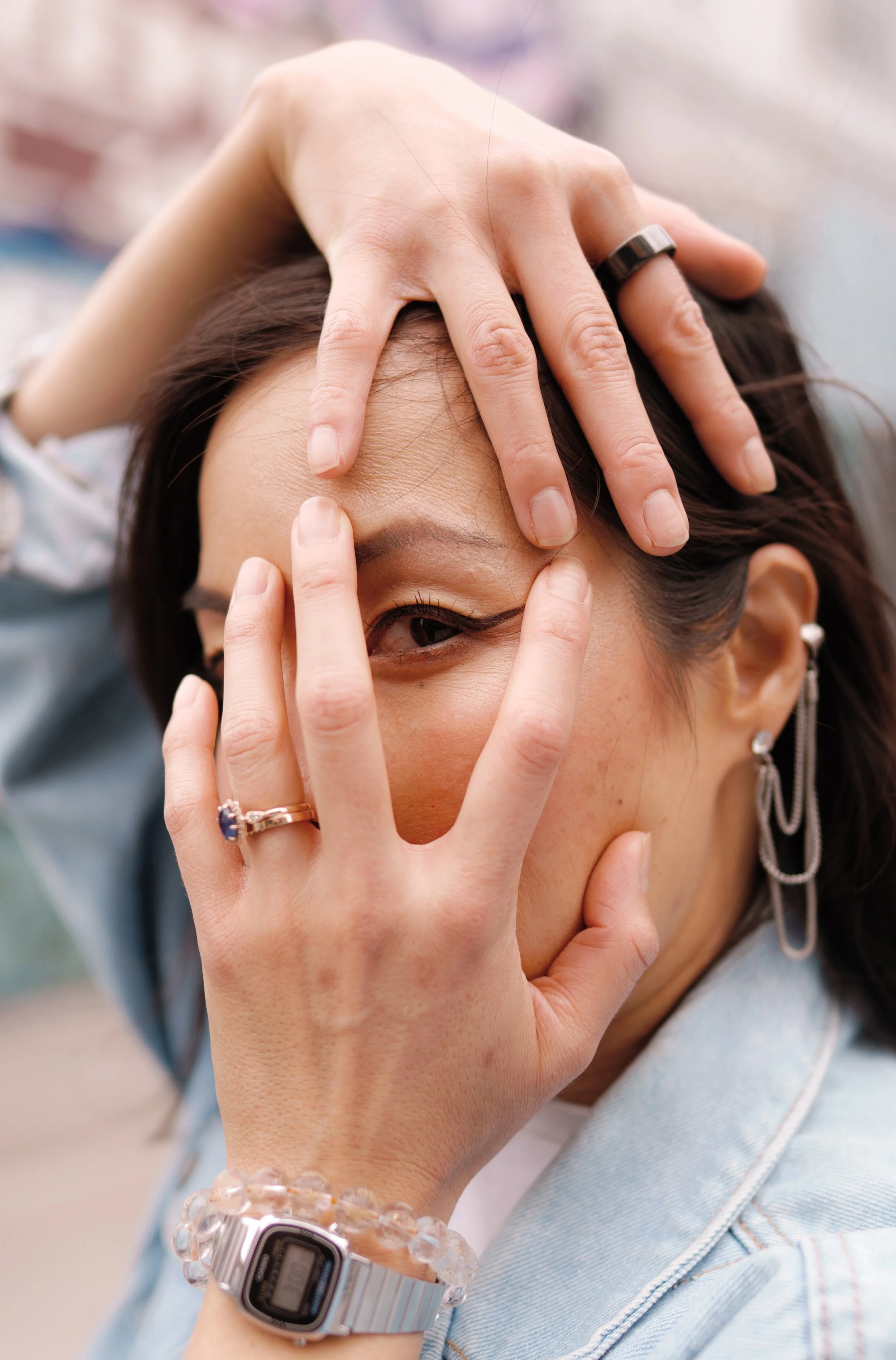 A person with dark hair holding their face with one hand, revealing their eye and part of their forehead. The person is wearing a silver wristwatch, a beaded bracelet, a ring, and earrings, and has makeup emphasizing their eye. The background is blur