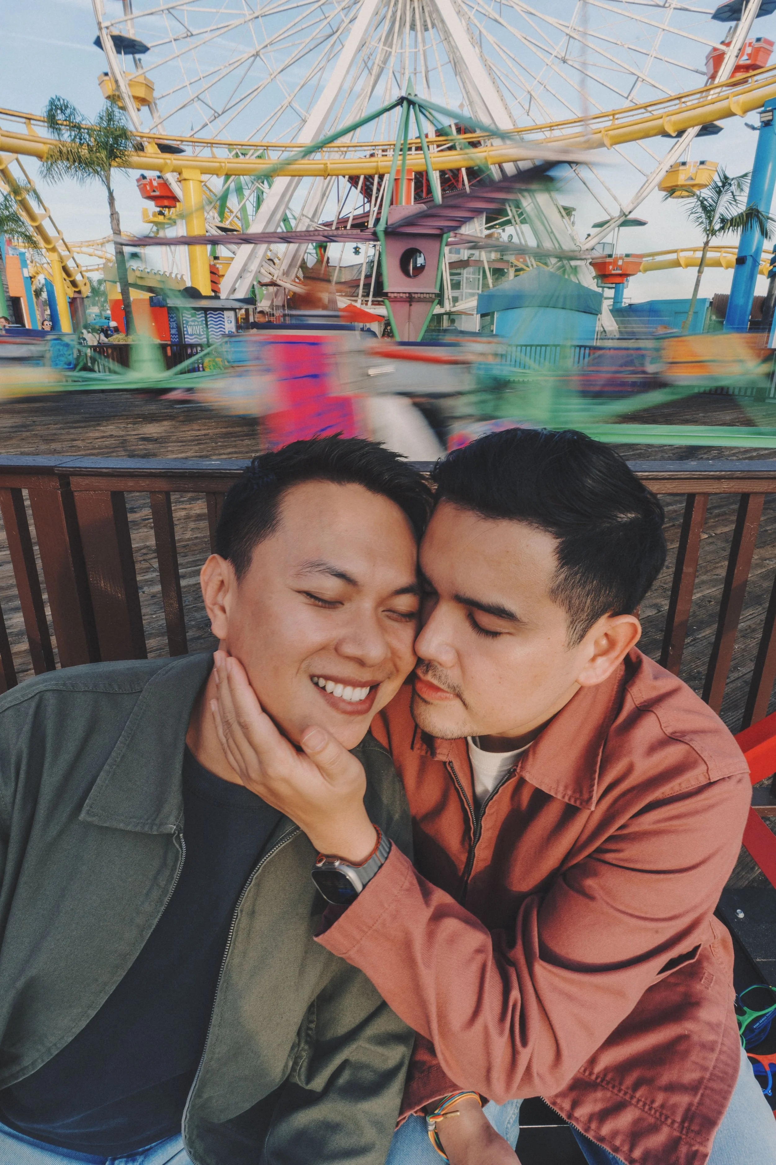 Two young men are smiling and cuddling in front of a moving amusement park ride with a Ferris wheel in the background.