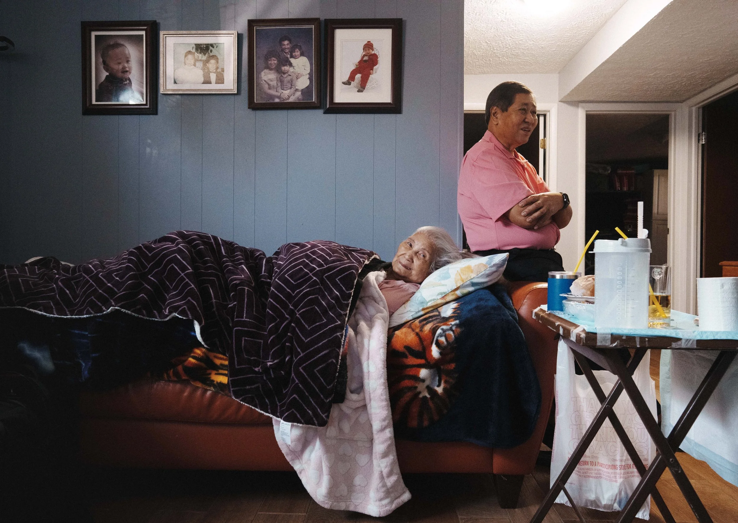 A woman lying on a sofa, an elderly woman lying in bed, and a man standing beside the bed in a living room. The wall has framed family photos. There is a table with drinks and a container, and a folded chair nearby.