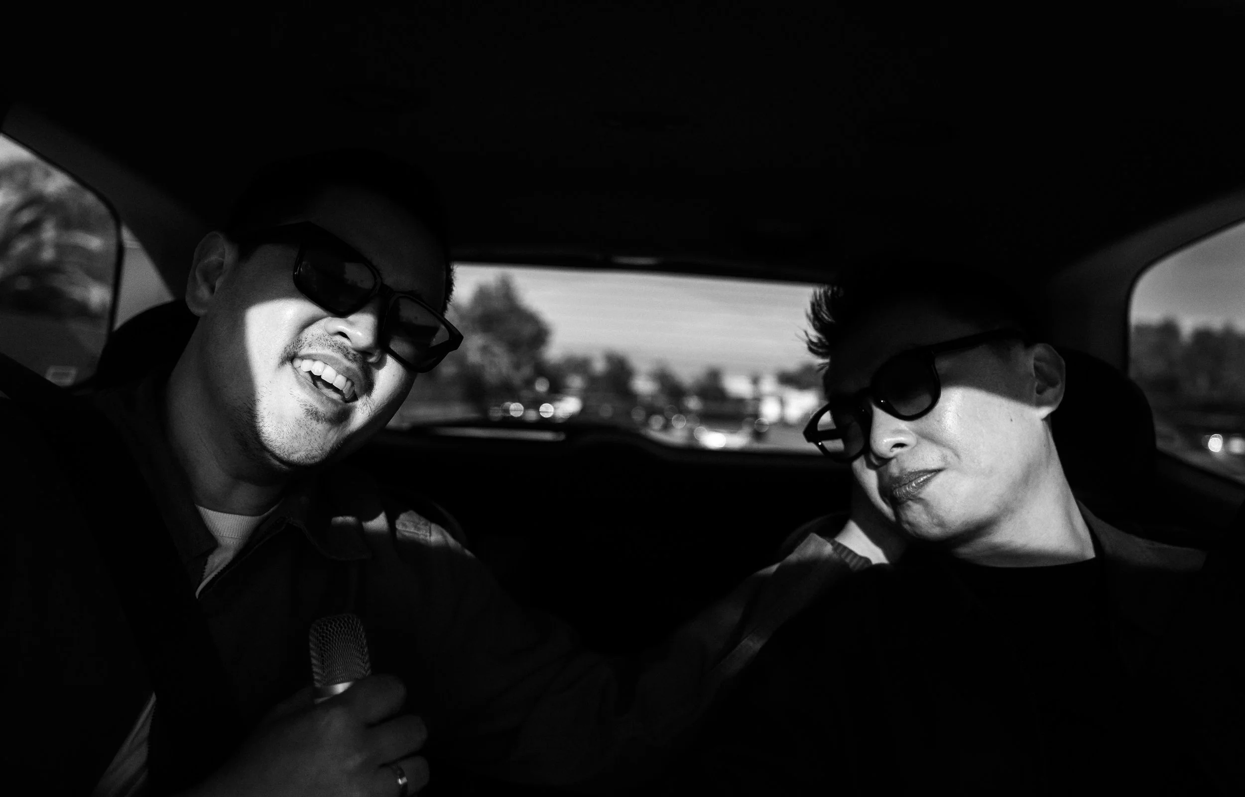 Two people wearing sunglasses sitting in a vehicle, with one smiling and holding a microphone.