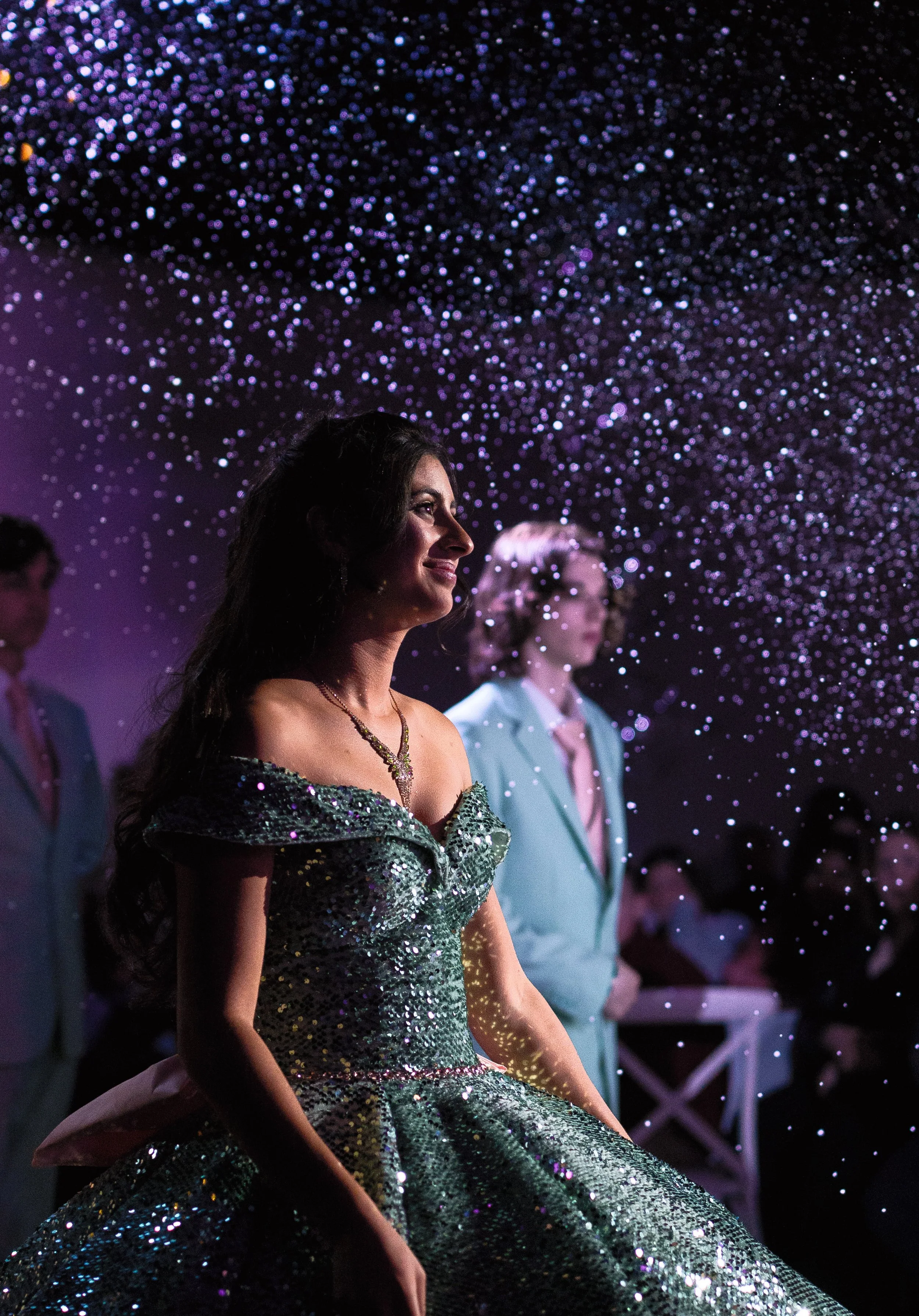 A woman in an off-shoulder silver gown sitting with a slight smile during a formal event, with a woman in a light green suit in the background and purple confetti or lights overhead.