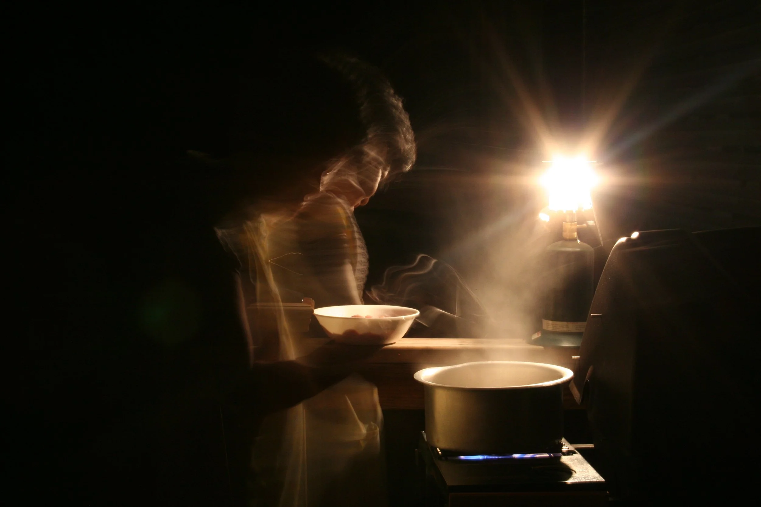 Person cooking at a stove illuminated by a bright lamp in a dark room, with bowls and utensils on a counter.
