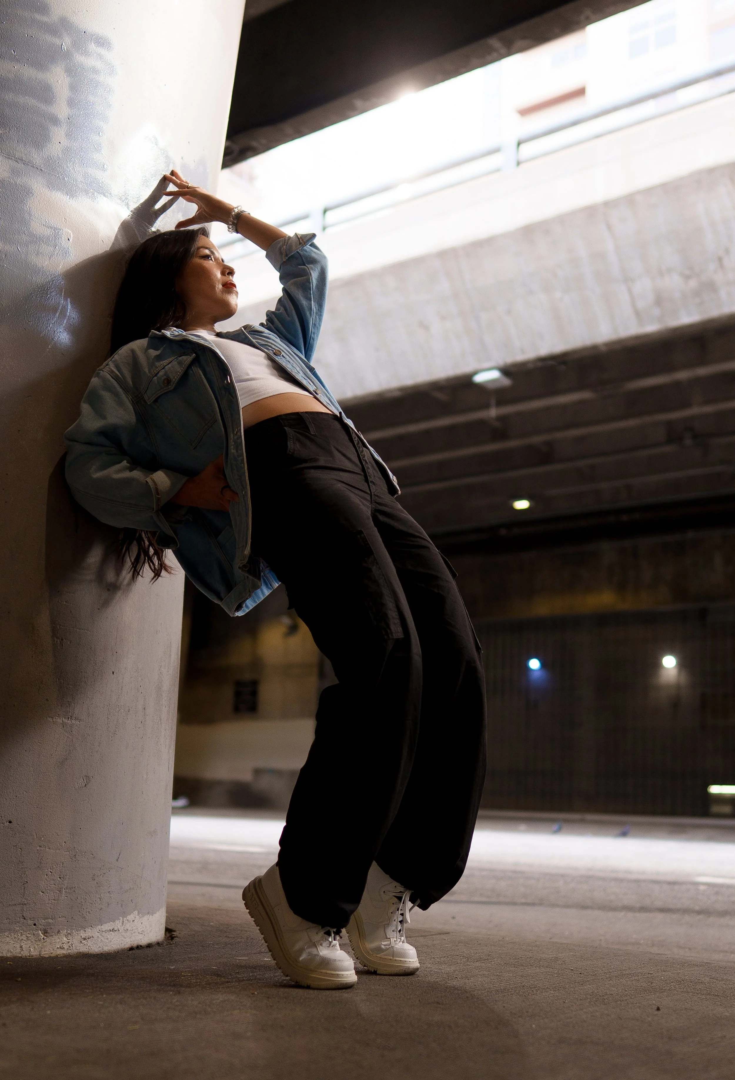 A woman leaning against a concrete pillar under an overpass, wearing a denim jacket, black pants, and white sneakers, with her hand on her forehead and gazing into the distance.