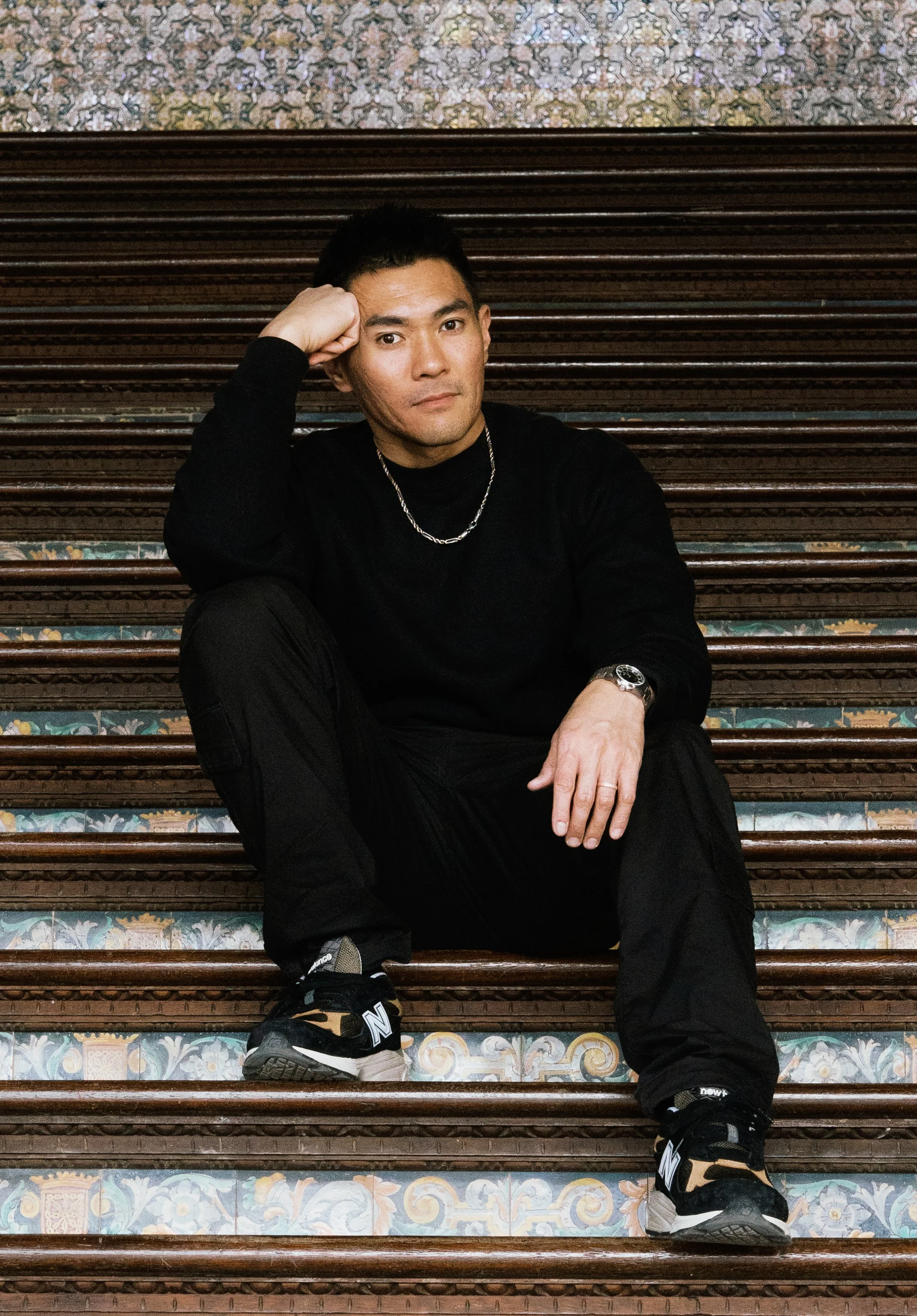 A man in black clothing sitting on a set of ornate, colorful tiled stairs, resting his head on his hand with a relaxed expression.
