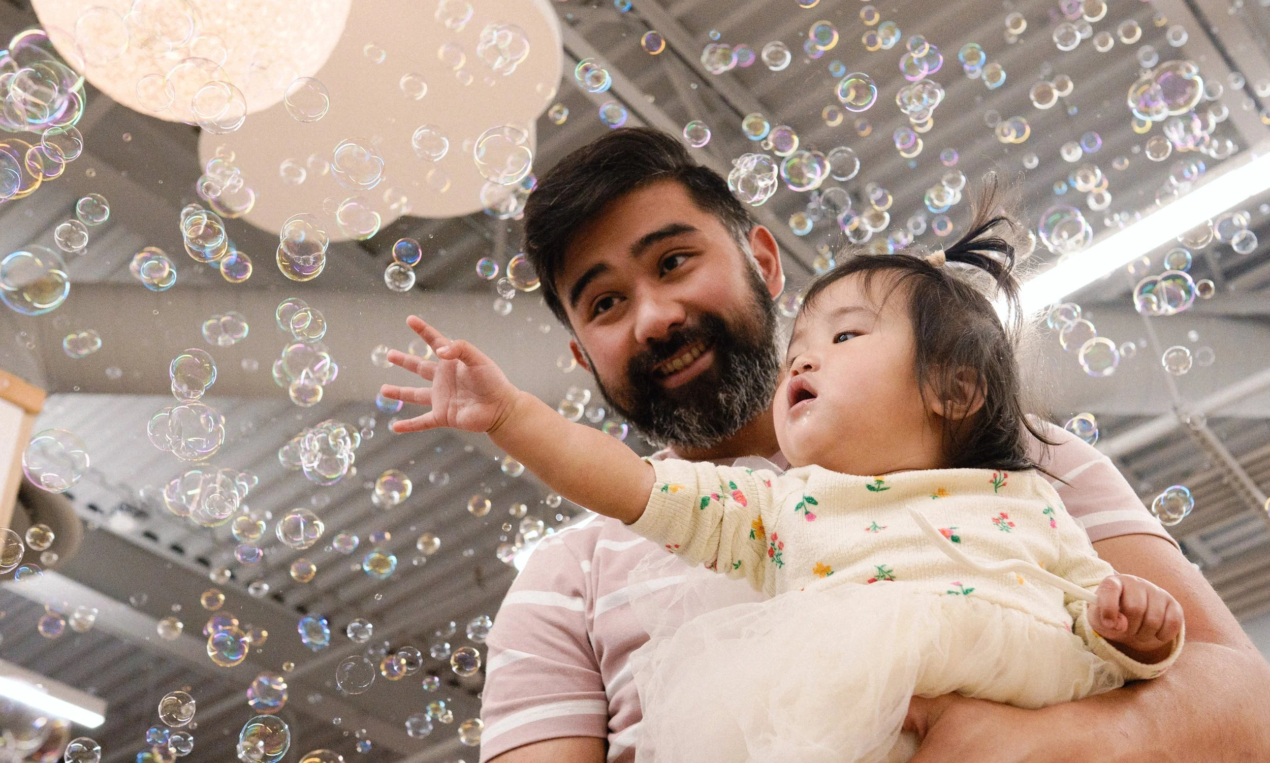 A man holding a young girl surrounded by floating soap bubbles indoors, with a metal ceiling above and bright lighting in the background.