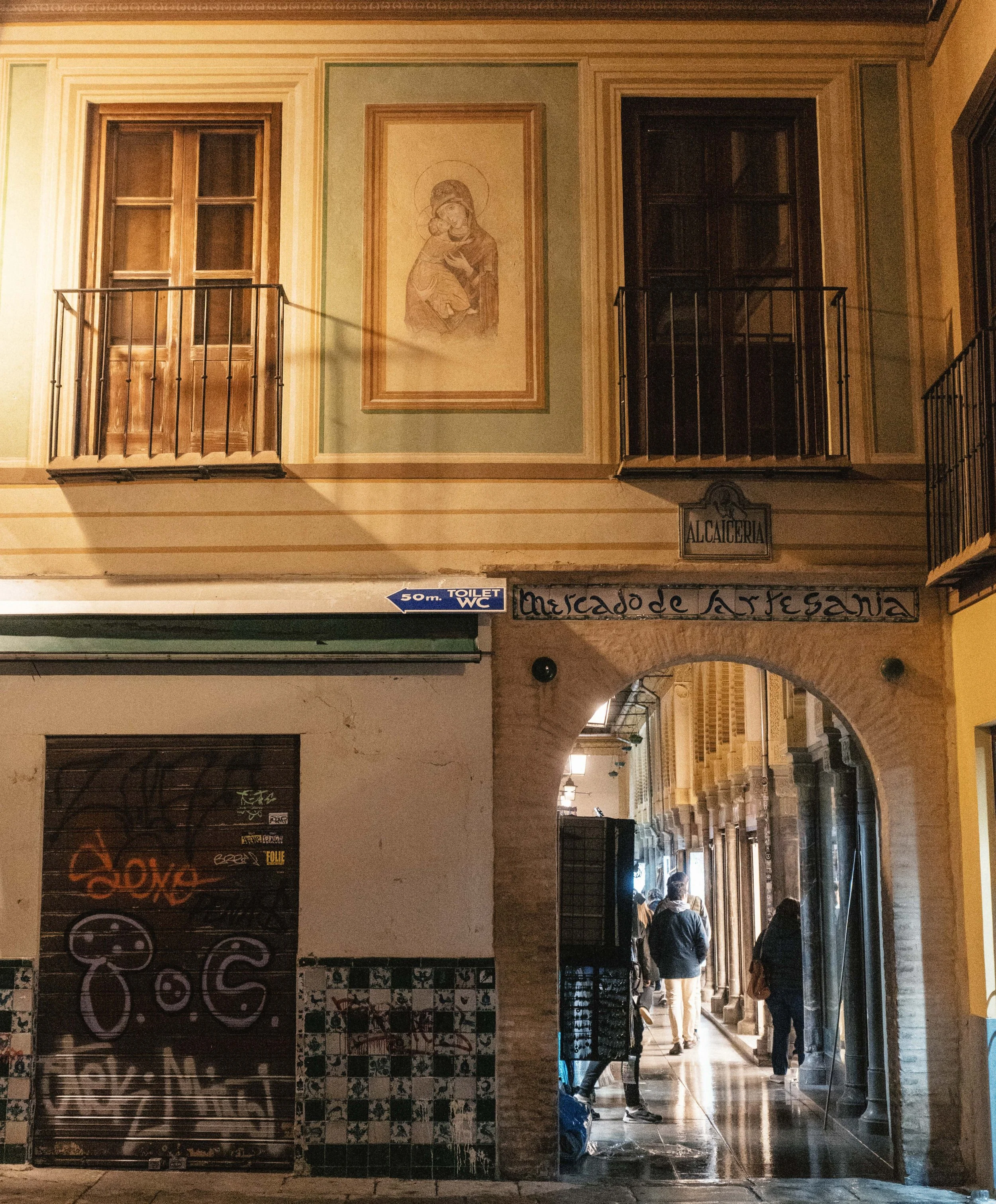 Night scene of a covered walkway or arcade with people standing in line, and a sign on the wall that reads 'Mercado de Santiago.' An archway is visible, and the street is illuminated with warm lighting.