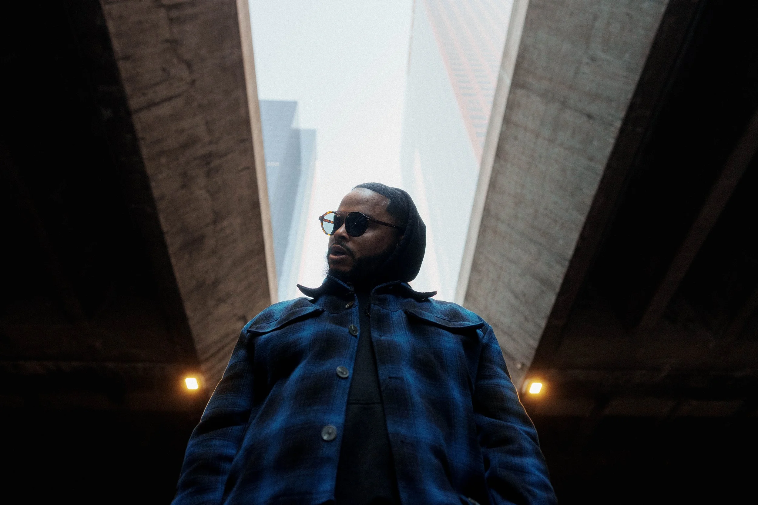A man wearing sunglasses and a hoodie standing under a bridge or overpass, looking slightly to the side, with tall buildings visible in the background.