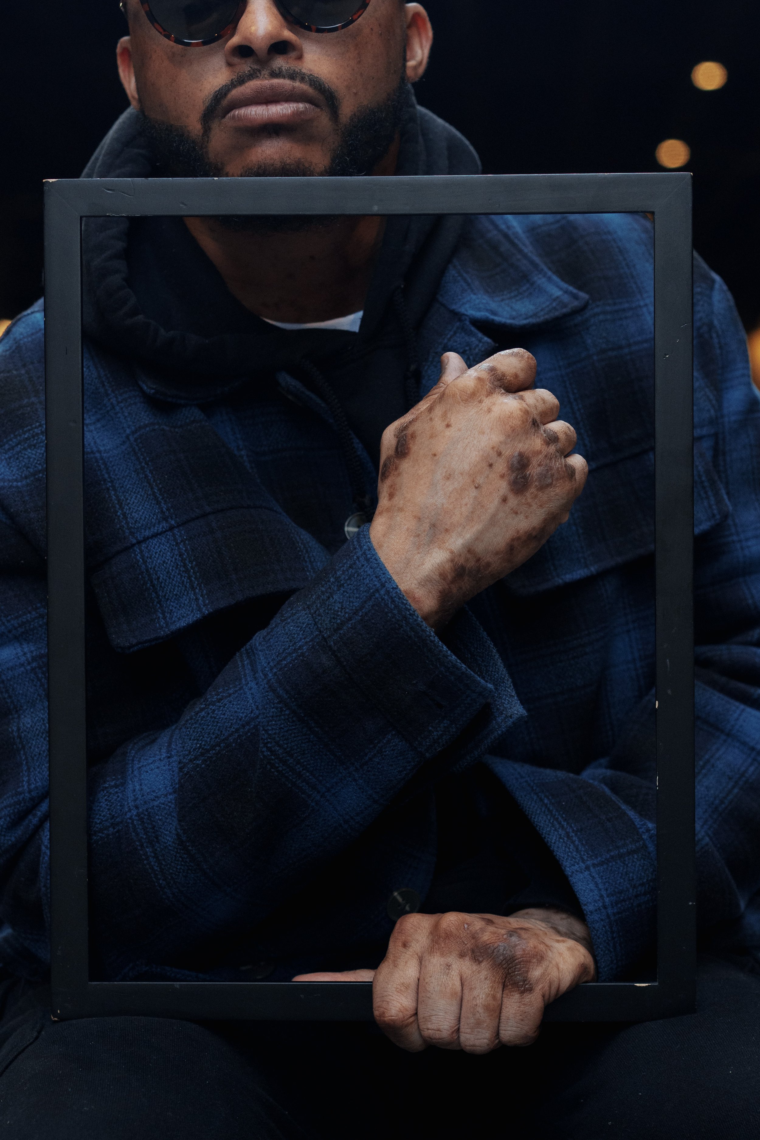 A man wearing sunglasses, a dark jacket, and a blue plaid shirt is holding a black picture frame in front of his face, with part of his face visible behind it. His hand appears aged with visible veins and wrinkles.