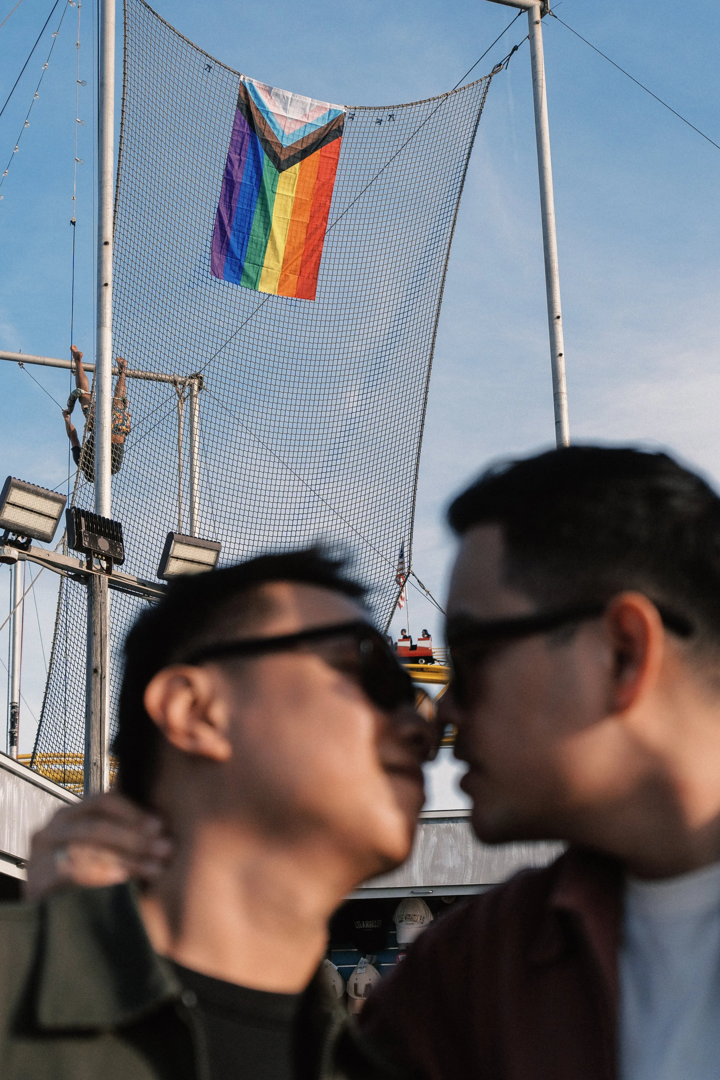 Two young people about to kiss at an amusement park, with a background of a climbing net and a flying rainbow flag. The photograph is taken outdoors during daytime.