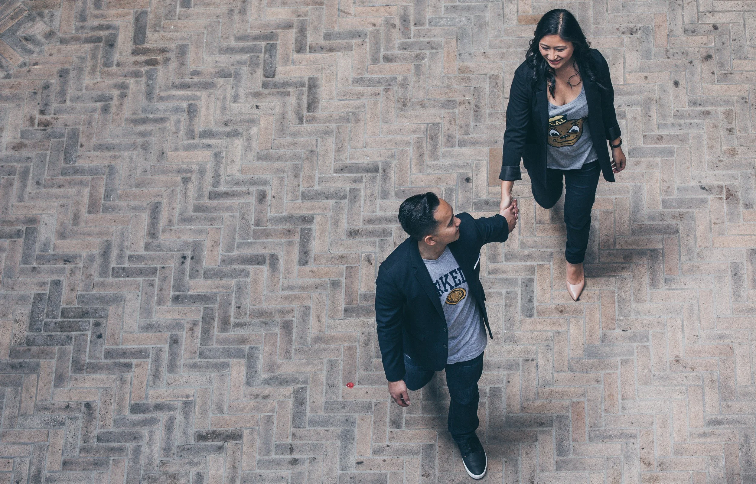 A woman and a man holding hands, walking on a paved brick surface, viewed from above. The woman is smiling, with black hair and wearing a blazer, jeans, and beige shoes, while the man has short dark hair and is dressed in a dark blazer and gray t-shi