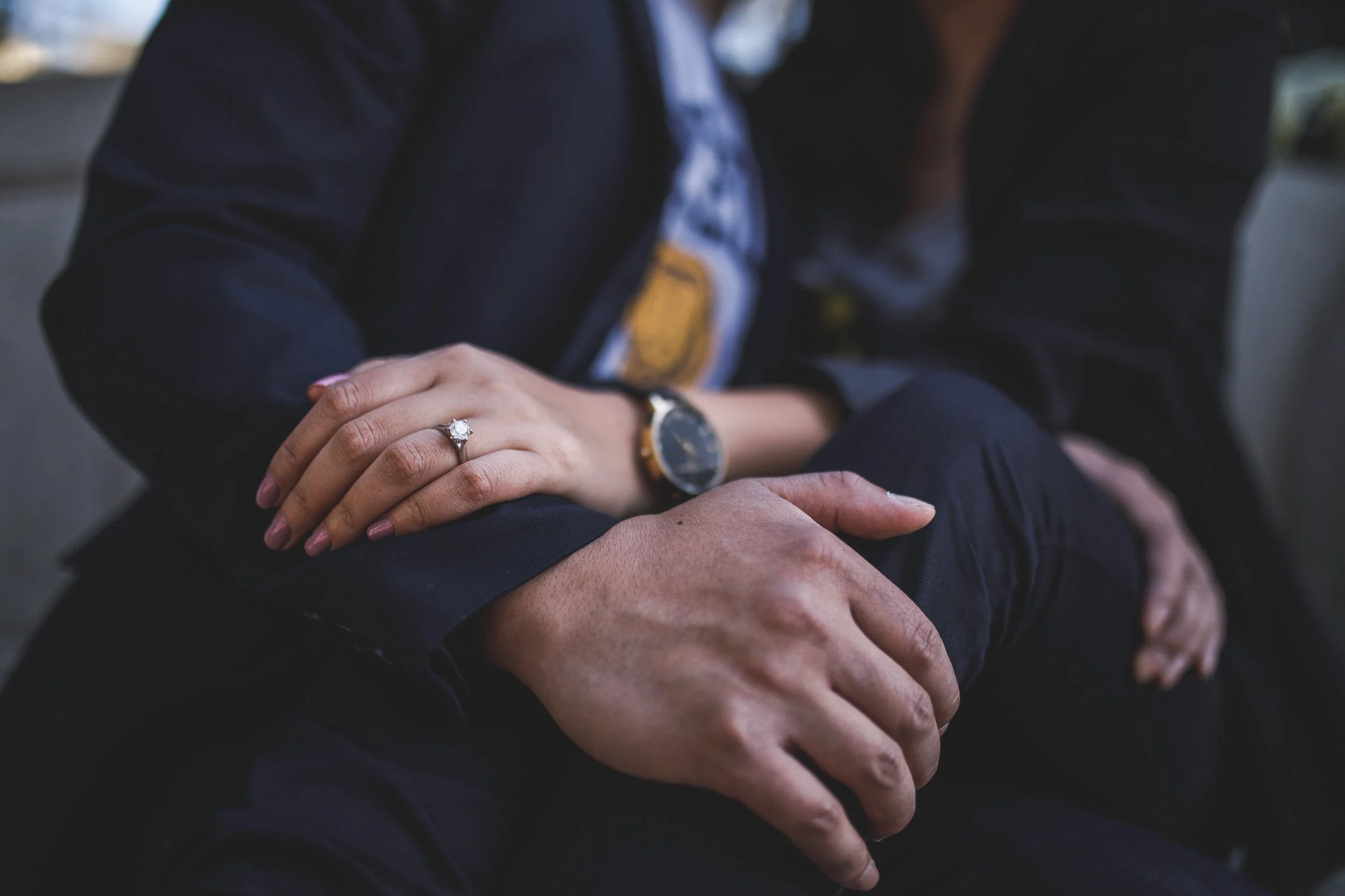 Close-up of a woman's hand wearing an engagement ring resting on another person's arm, with a blurred background.
