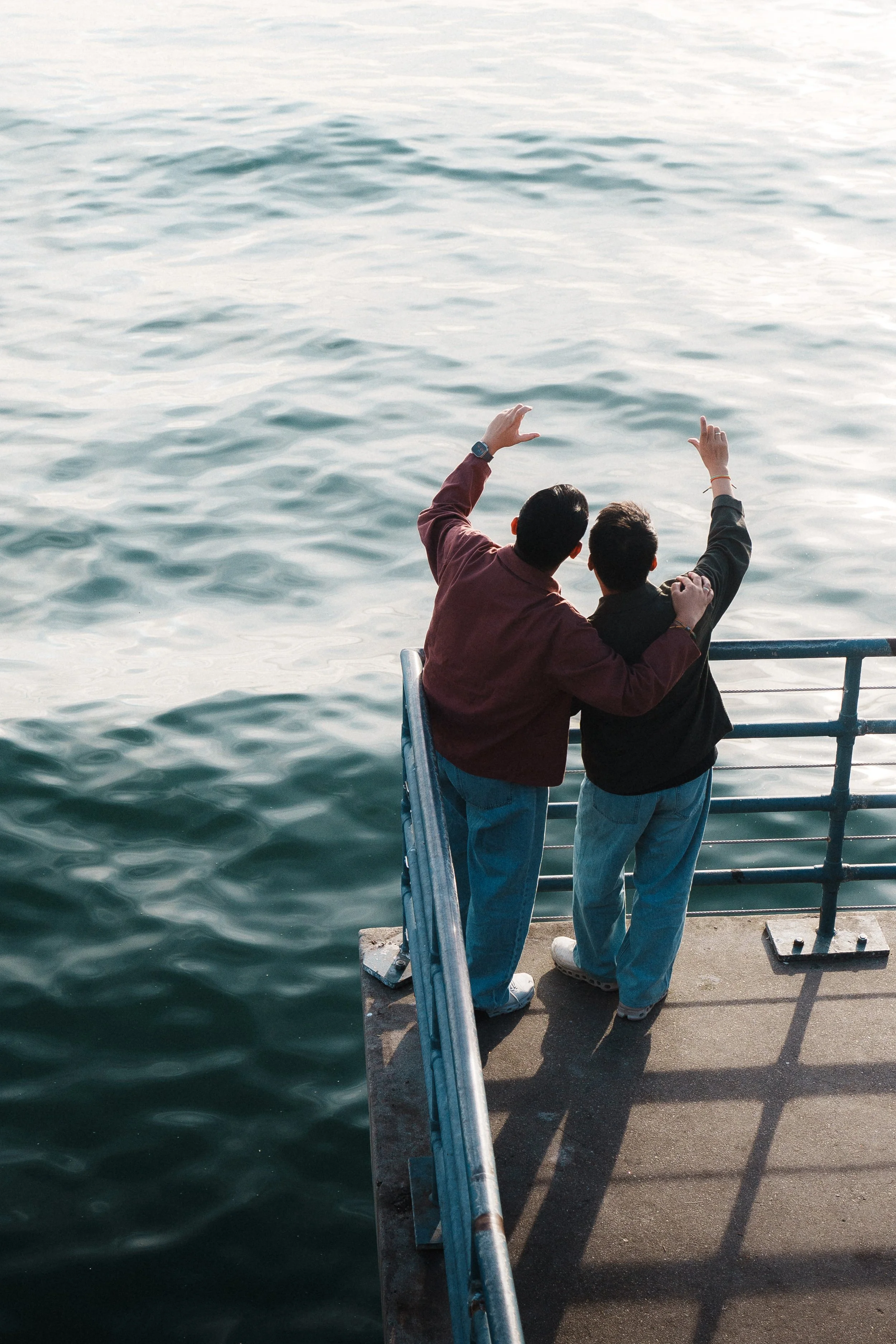 Two young men standing on a concrete pier, looking out at the water. They are leaning on a metal railing, with one of them raising his hand and pointing towards the water, while the other has his arm around his friend's shoulder. The water is calm wi