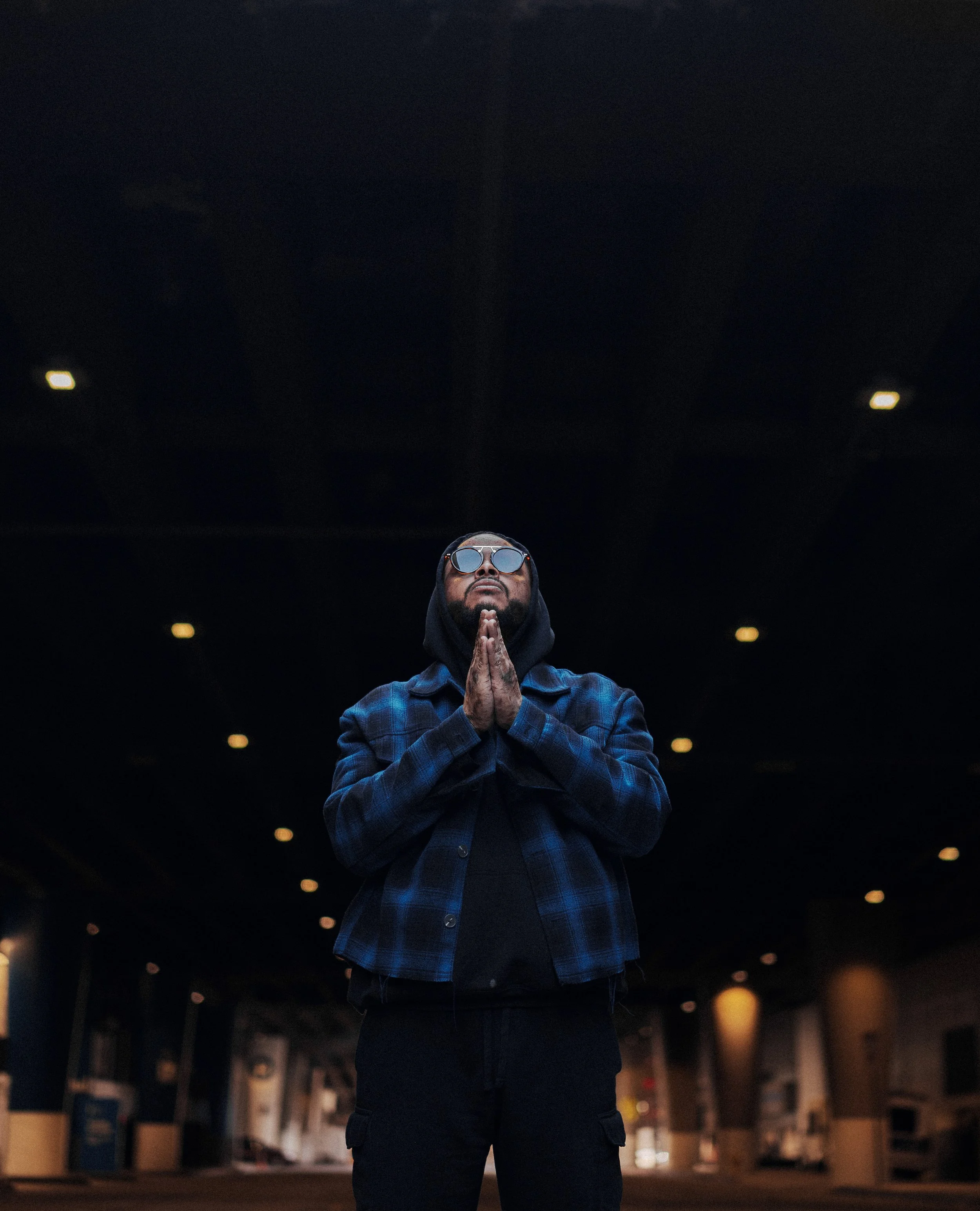 A person standing outside at night with their hands pressed together in a prayer or meditative pose, wearing reflective sunglasses, a blue plaid shirt, and a dark hoodie, with a dark building or parking garage in the background.
