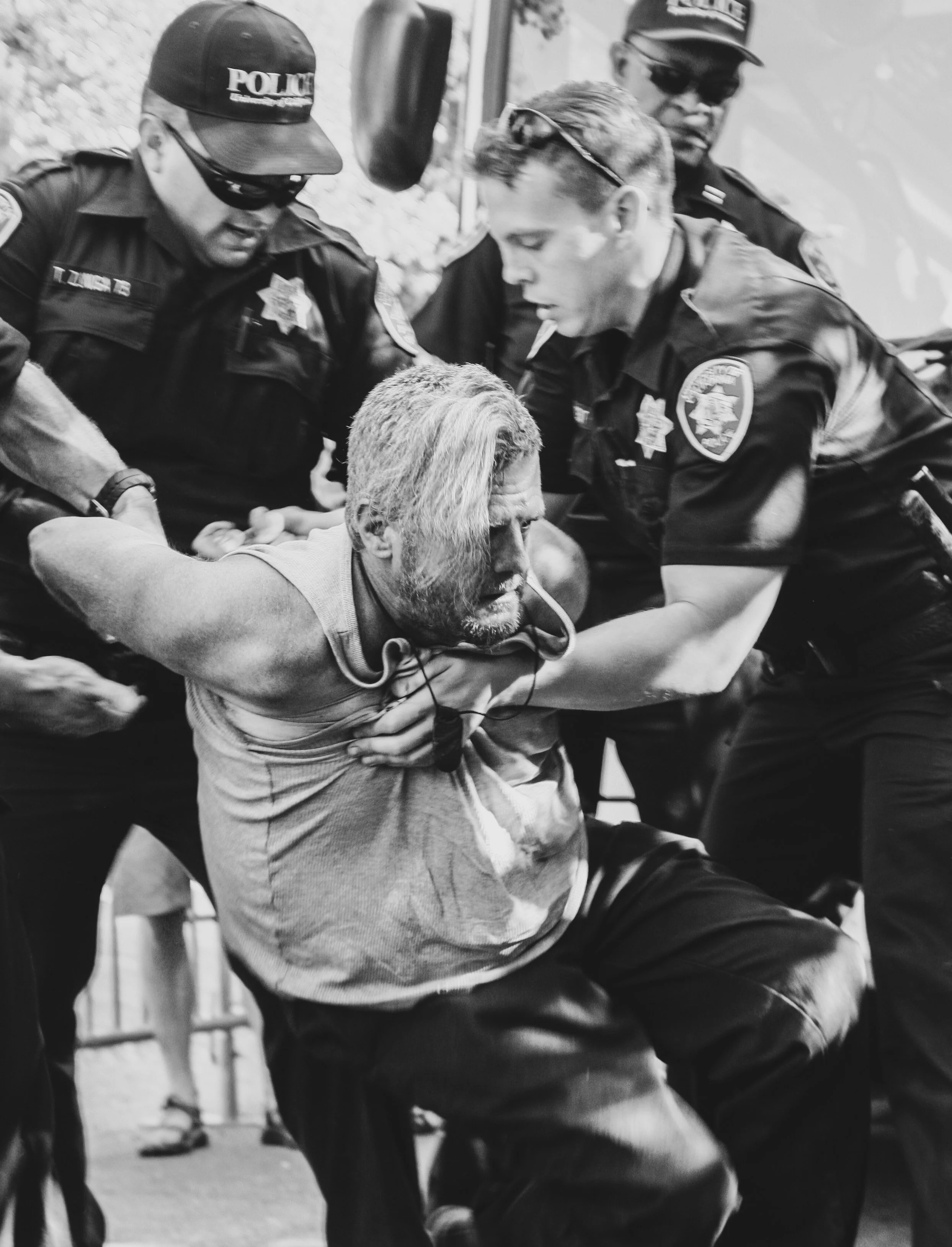 Black and white photo of police officers detaining an elderly man with grey hair and a beard, who appears to be held by the officers.
