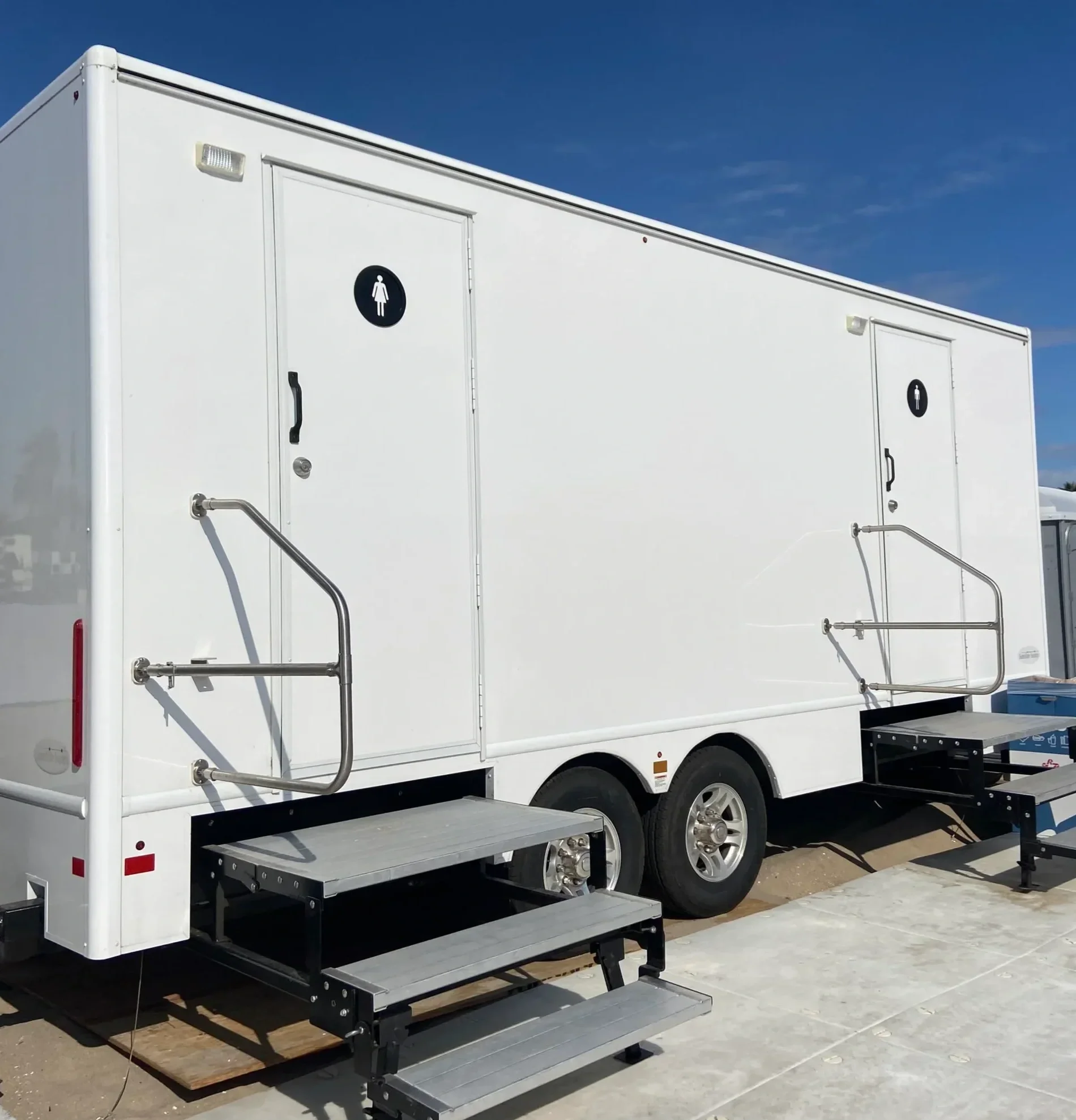White mobile restroom trailer with two doors marked with female symbols, outdoor setting, clear blue sky, black stairs and metal handrails at entrances.