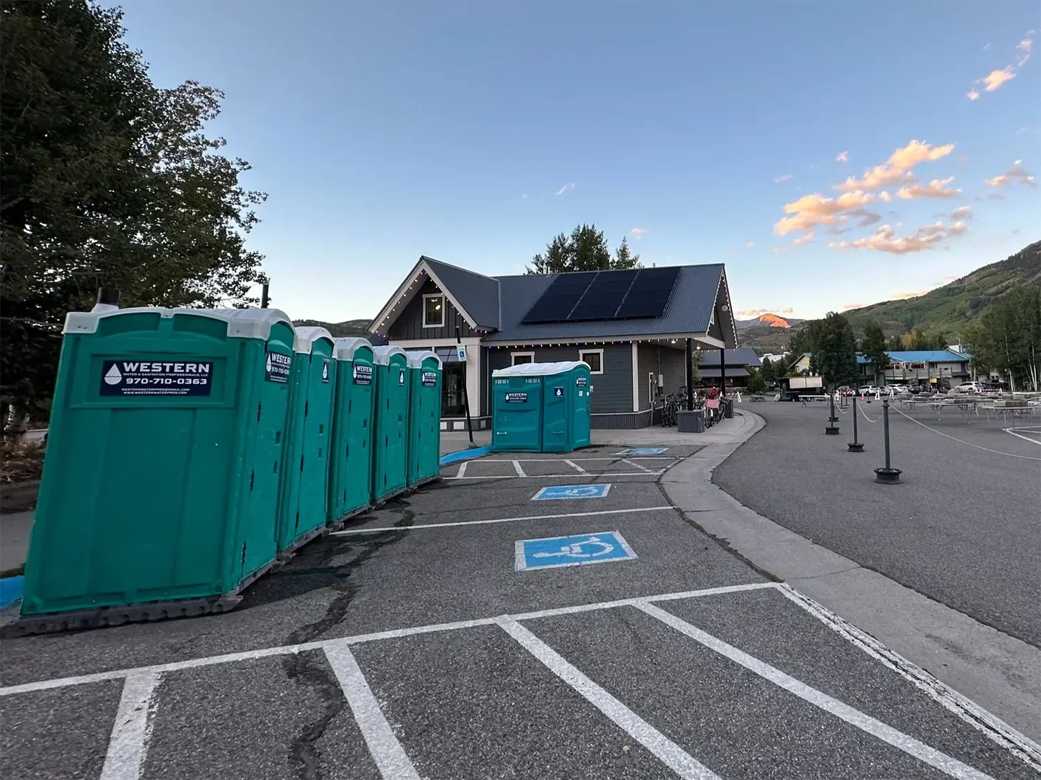 A row of portable toilets next to a building with solar panels and string lights, in an outdoor parking lot with handicapped parking spaces and a mountain view in the background at sunset.