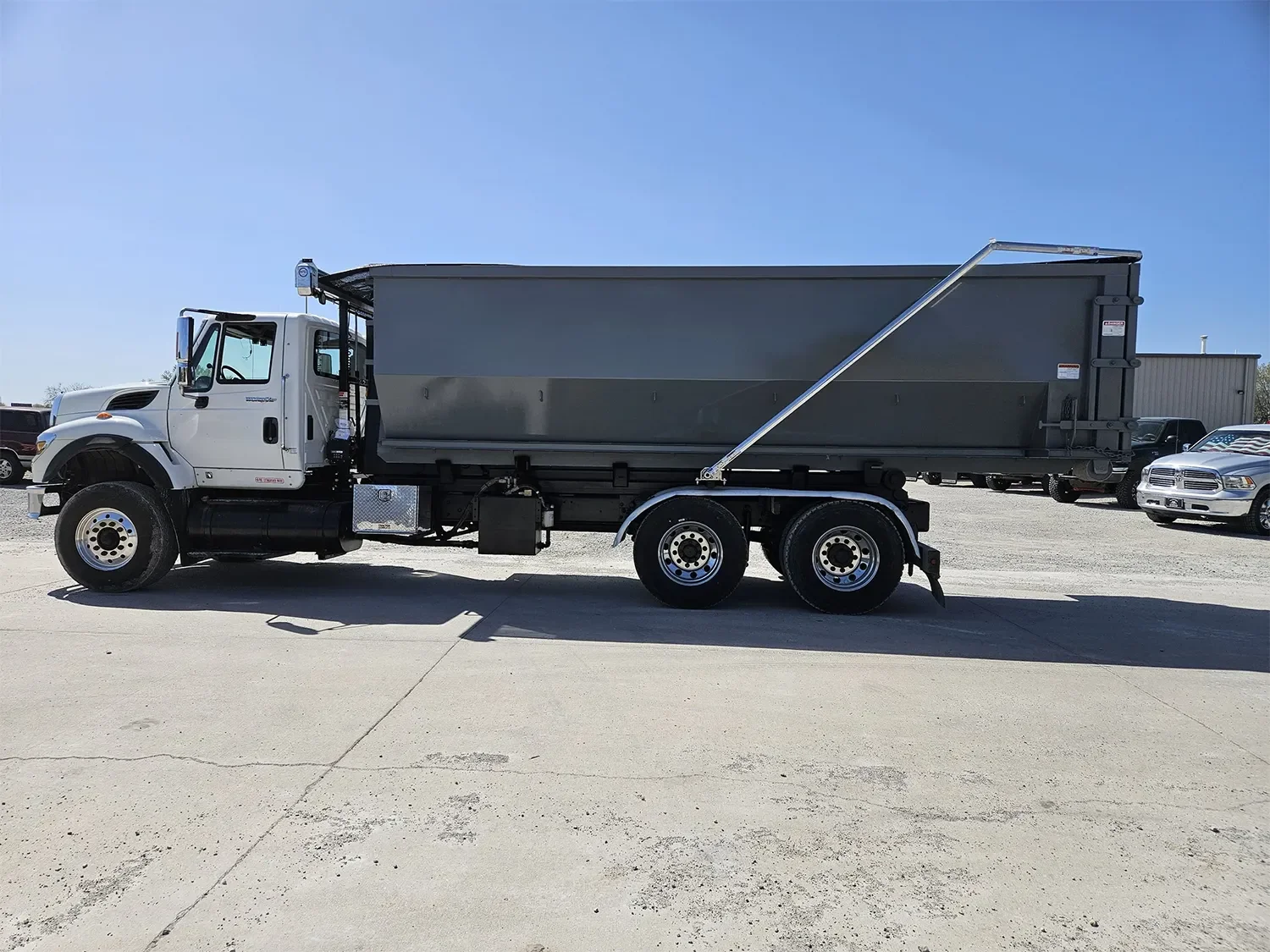 A white industrial dump truck with gray dump bed parked on a concrete lot under a clear blue sky. Several cars are visible in the background.