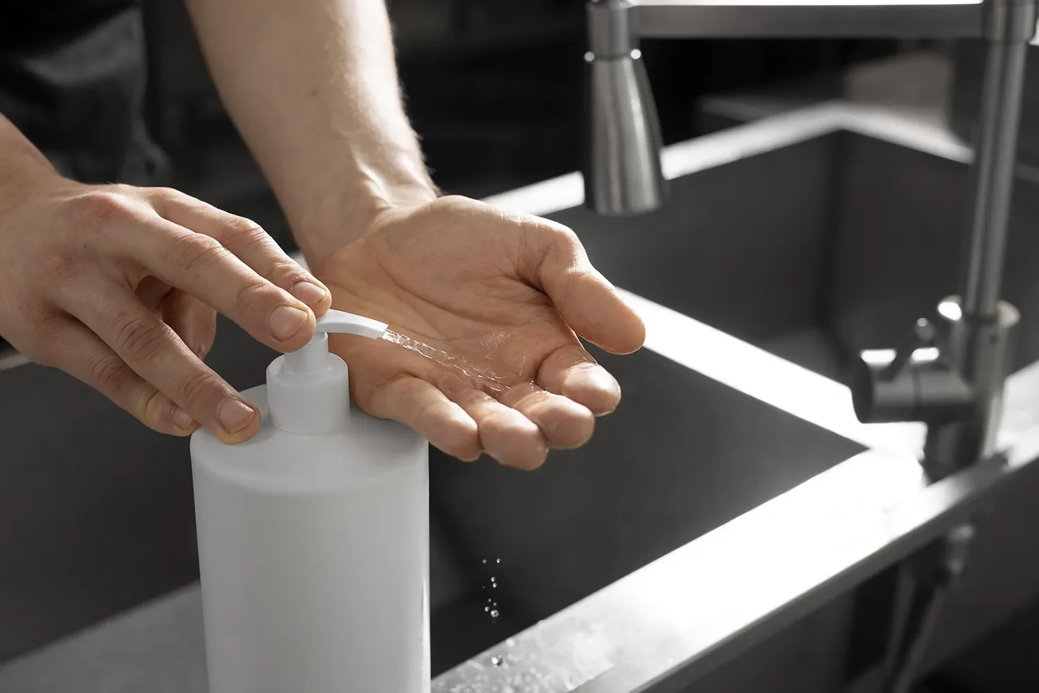 Person dispensing hand sanitizer from a white bottle onto their palm in a kitchen or sink area.