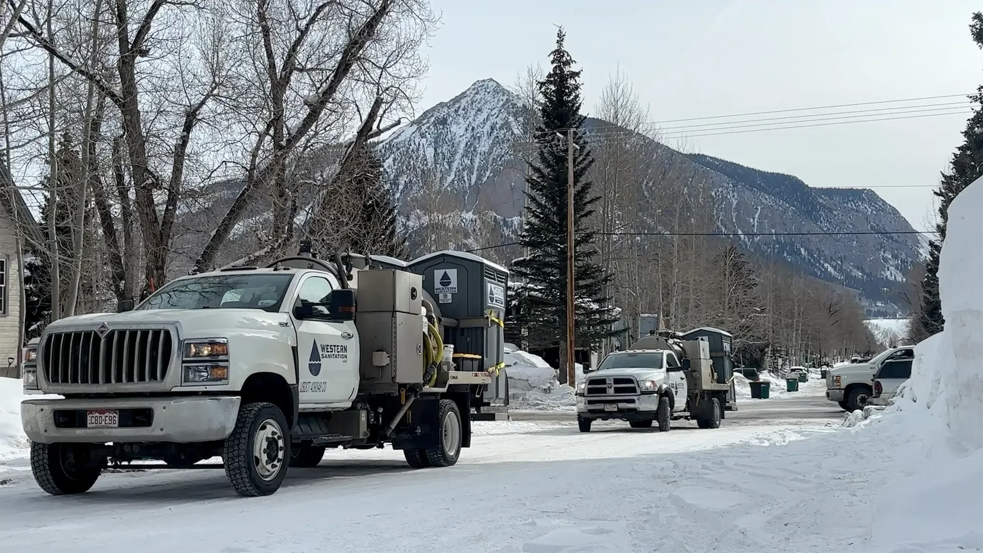 Street scene with trucks and utility vehicles parked on snow-covered road, with snow-capped mountain and leafless trees in the background.