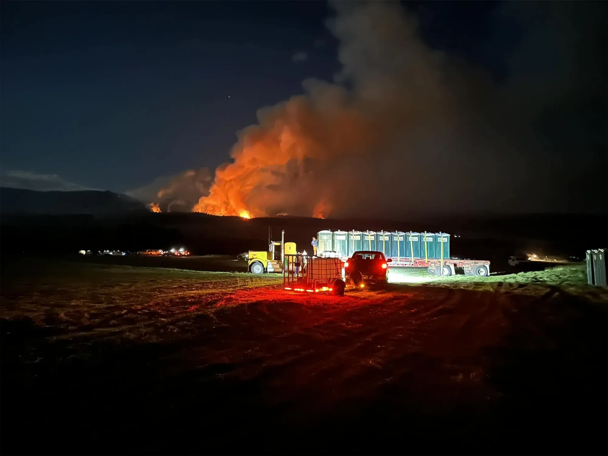 Nighttime scene with fire on distant hills, large smoke plume rising into dark sky, and trucks with bright lights parked on grassy area in foreground.
