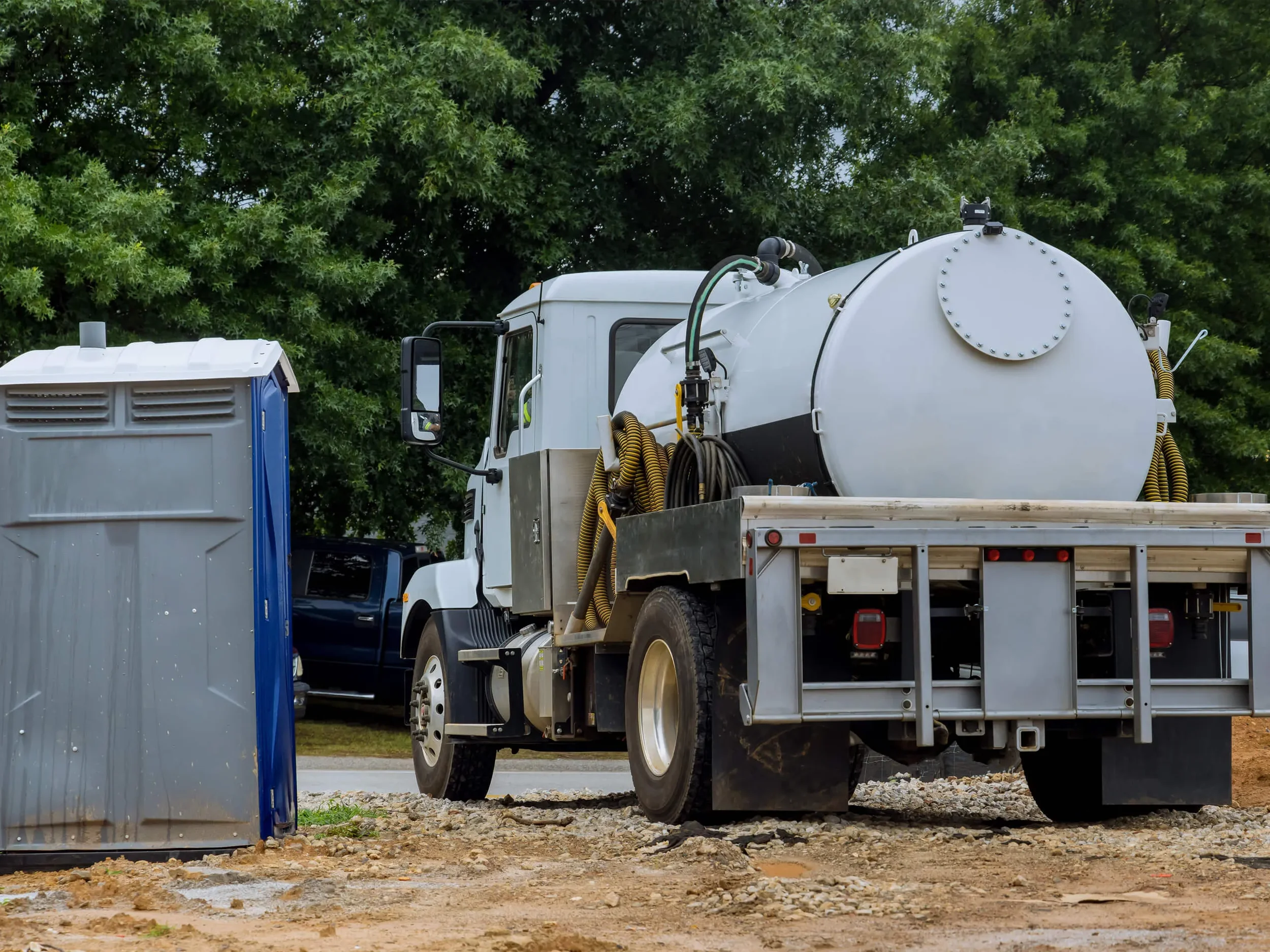 A street sweeper truck with a large water tank and hoses near a construction site, with trees in the background.