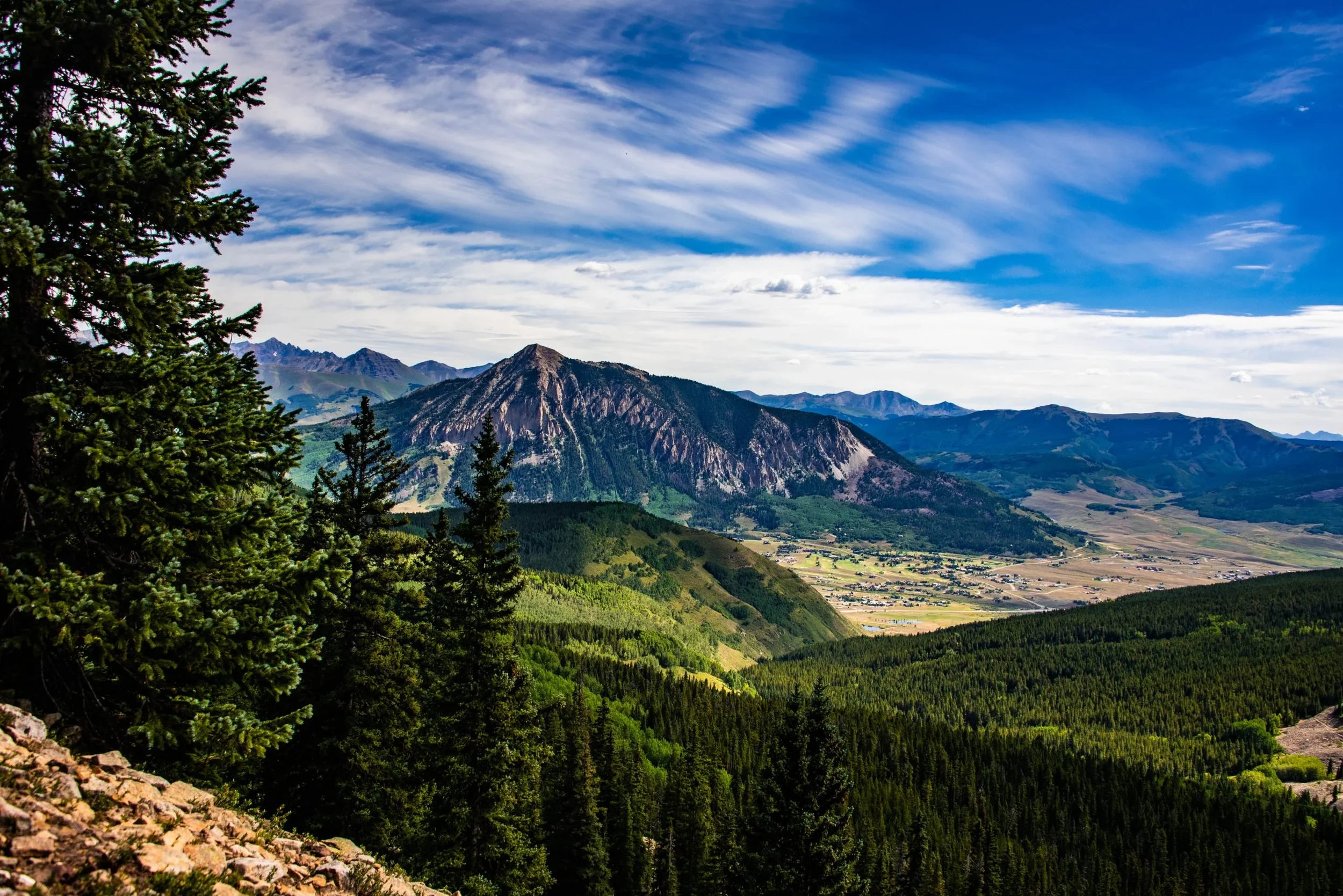 A scenic mountain landscape with lush green forests, rocky peaks, and scattered farmland under a partly cloudy sky.