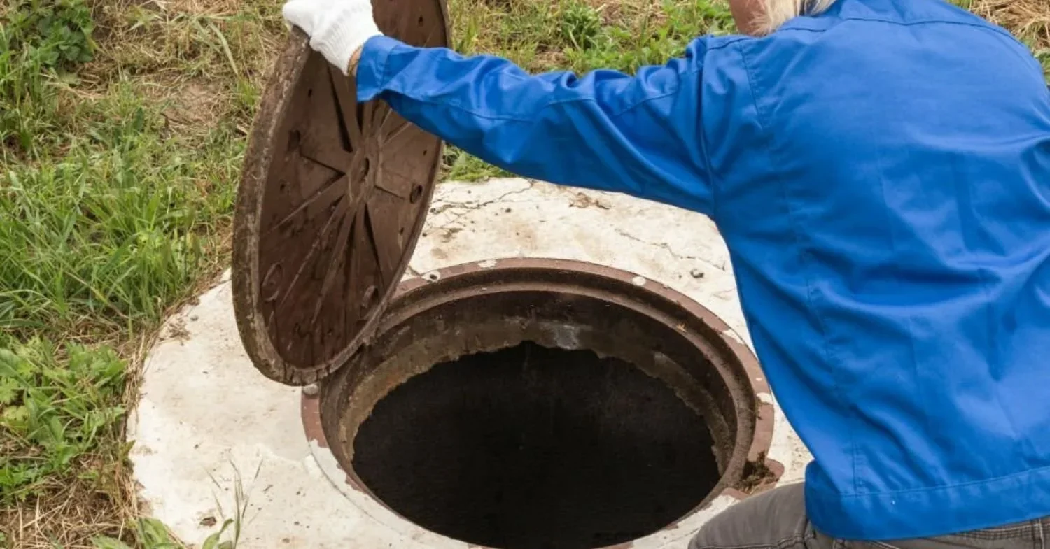 A person in a blue jacket and white gloves opening a manhole cover to reveal a dark underground space.