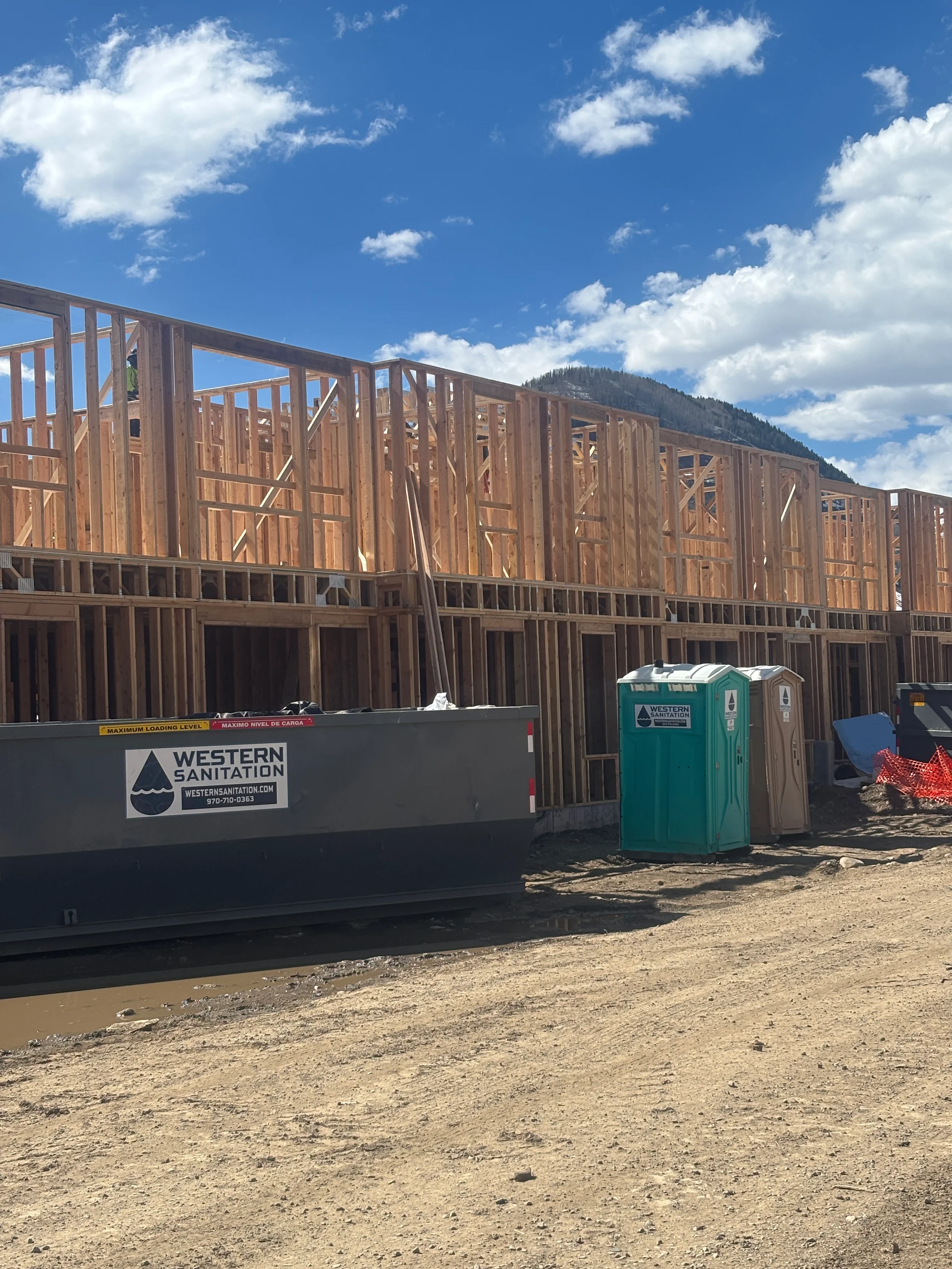 Construction site with wooden framework for a building, portable toilets, and a dumpster with Western Sanitation branding. Mountain in background and partly cloudy sky.