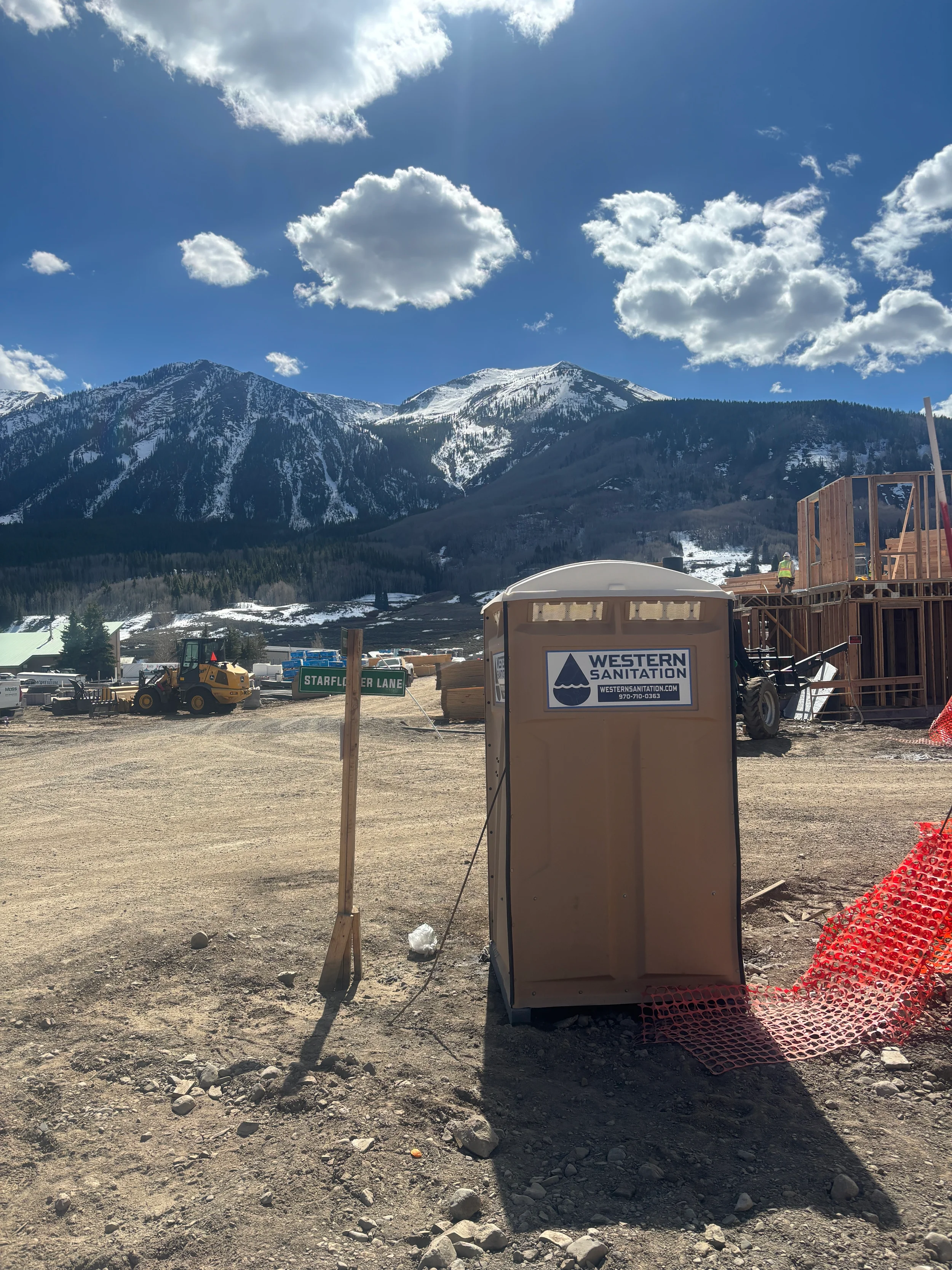 A portable toilet with a sign that says 'Western Sanitation' at a construction site with mountains in the background, blue sky, and clouds.