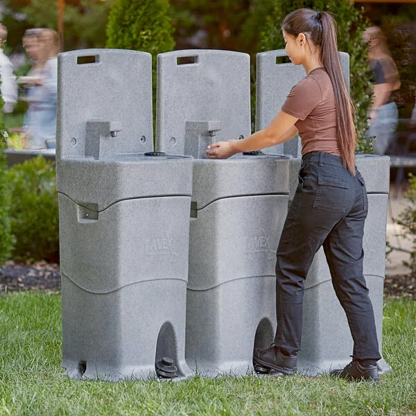 A woman uses a gray, modular, portable drinking water station outdoors on a grassy area, with trees and people in the background.