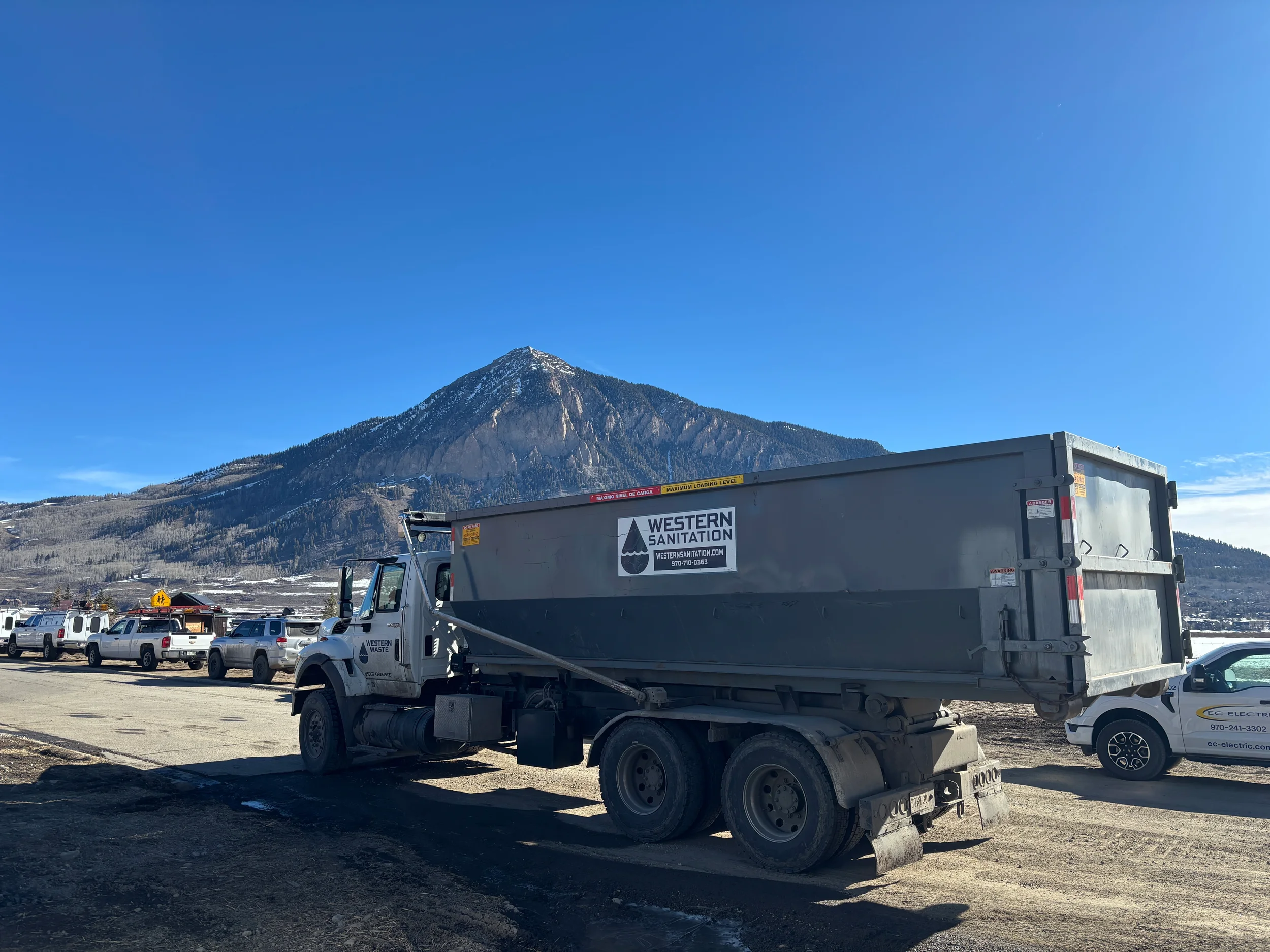 A large waste disposal truck with Western Sanitation branding parked in a lot with other vehicles, against a backdrop of a mountain with some snow and a clear blue sky.