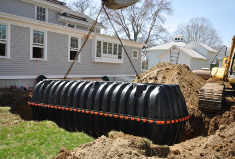 A large black septic tank being lowered into an excavated trench by a crane at a backyard construction site.