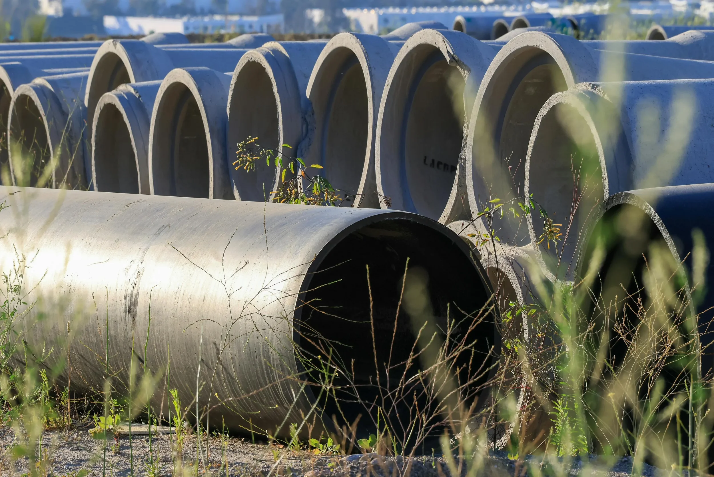 Large concrete pipes stacked outdoors with grass and weeds growing around them.
