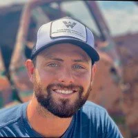 A smiling man with a beard wearing a blue shirt and a white and grey cap, outdoors with a blurry background of trees and rusty machinery.